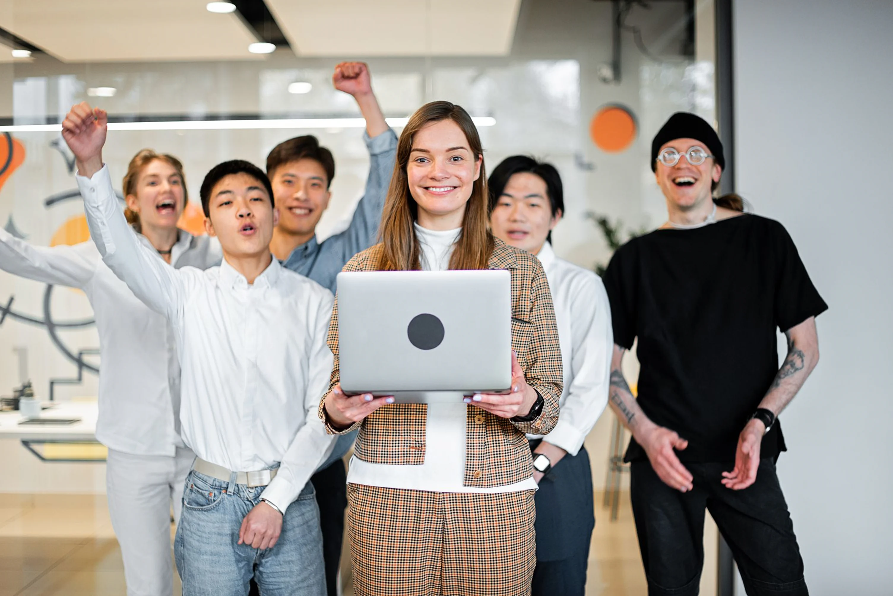 A diverse group of young professionals celebrates together in a modern office environment. The woman in the center smiles warmly while holding a closed laptop, as her colleagues cheer enthusiastically behind her.