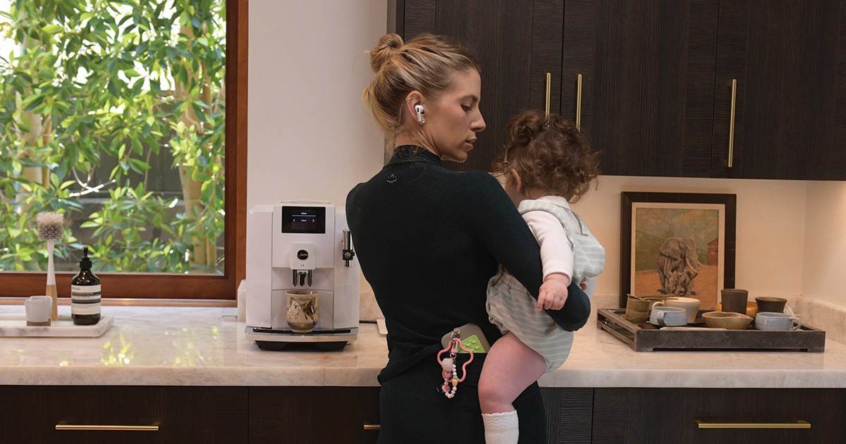 Woman holding a child in a kitchen while listening through earbuds during a busy everyday moment.