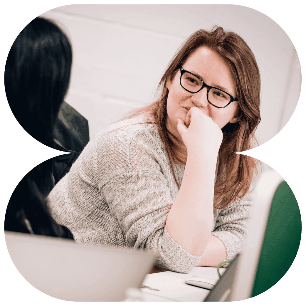 Person seated at a desk leaning on a hand while listening to another person beside a laptop.