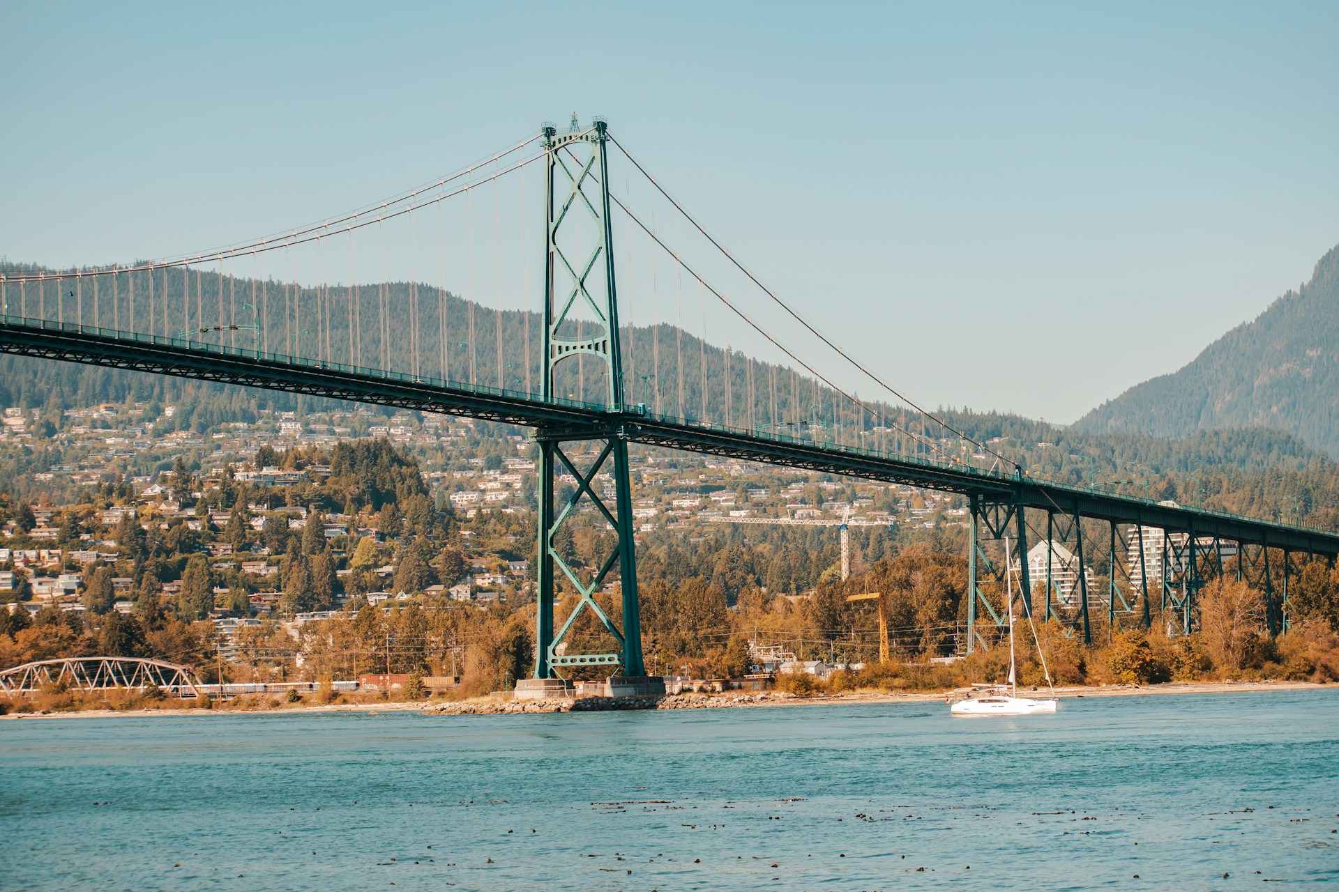 A green suspension bridge spanning across a body of water, with forested hills and residential homes in the background.