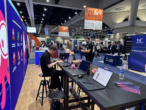 A person with short, dark hair, wearing a black long-sleeve shirt, stands against a dark background with their arms crossed and a neutral expression.