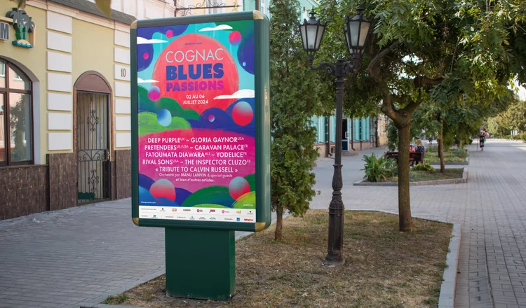 An outdoor sidewalk scene with a colorful billboard advertising a music event called 'Cognac Blues Passions' taking place from August 2 to July 6, 2024. The billboard lists performers such as Deep Purple UK, Gloria Gaynor USA, Pretenders UK/USA, Cara