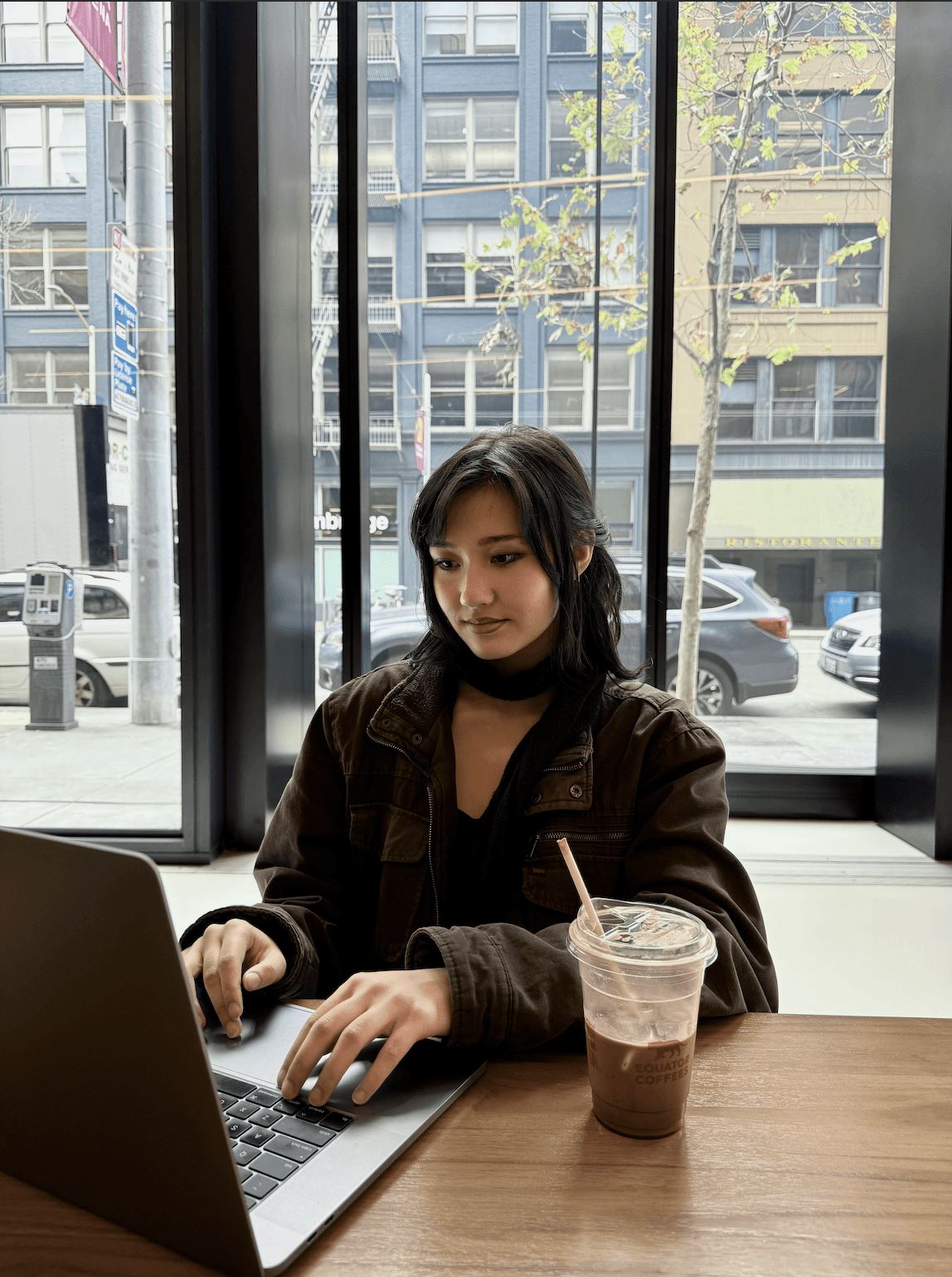 A young person with curly hair smiles at the camera while sitting in front of a laptop against a dark background.