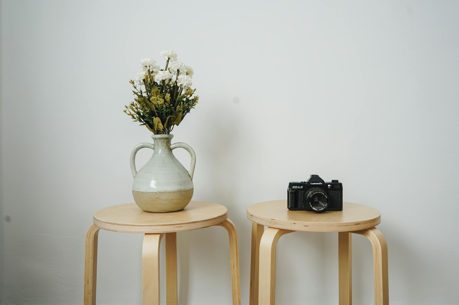 Two wooden stools beside a white wall, one holding a vase with flowers and the other a camera.