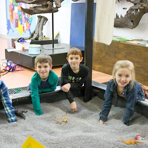 เด็กสี่คนกำลังเล่นในกระบะทรายที่มีของเล่นไดโนเสาร์ภายในนิทรรศการของพิพิธภัณฑ์