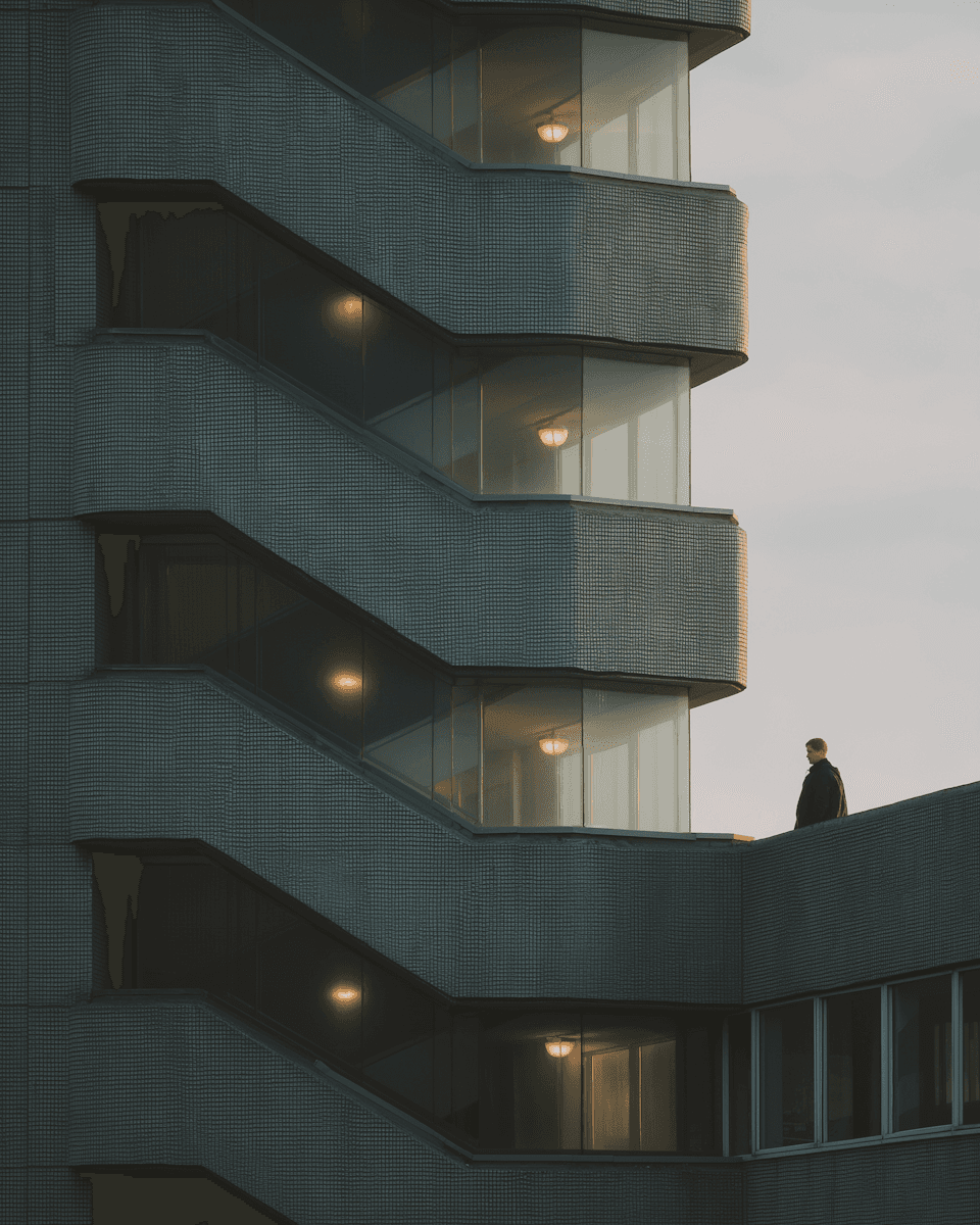 Modern building with staggered balconies, glass walls, and warm interior lighting. A person stands on a balcony, adding a human touch.
