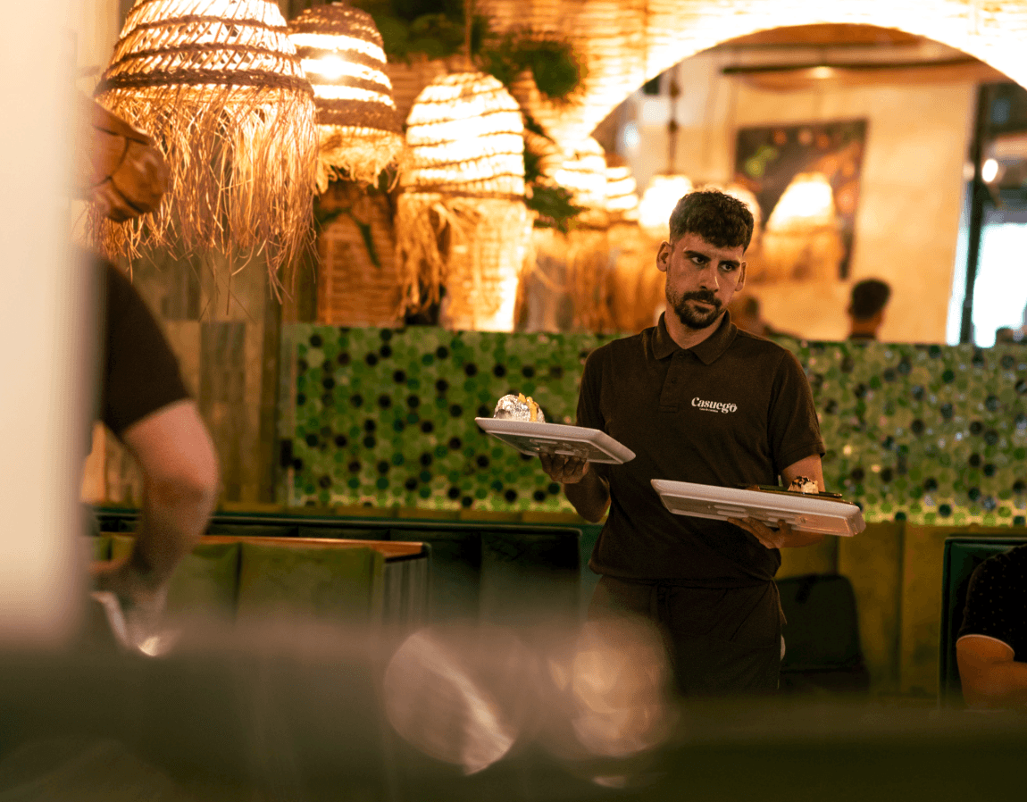 A serene and elegant spa space with wooden reclining chairs and white cushions. Golden sunlight filters through a cascading water feature, creating a peaceful atmosphere. Tropical plants and candles add to the relaxing ambiance.