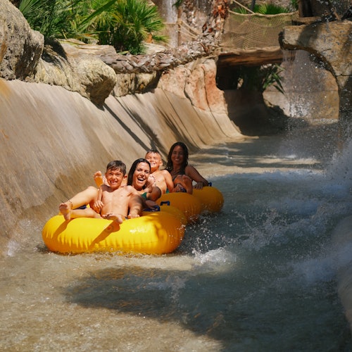 Four people smiling on a yellow inflatable raft, riding through a water slide surrounded by rocky terrain and lush greenery.