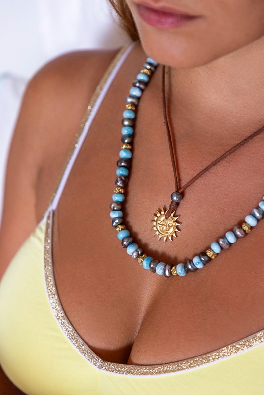 Handcrafted luxury jewelry by Manos de Oro Saint Barth—featuring a golden sun pendant paired with turquoise-toned and gold-detailed beads, expressing joyful Caribbean sophistication.