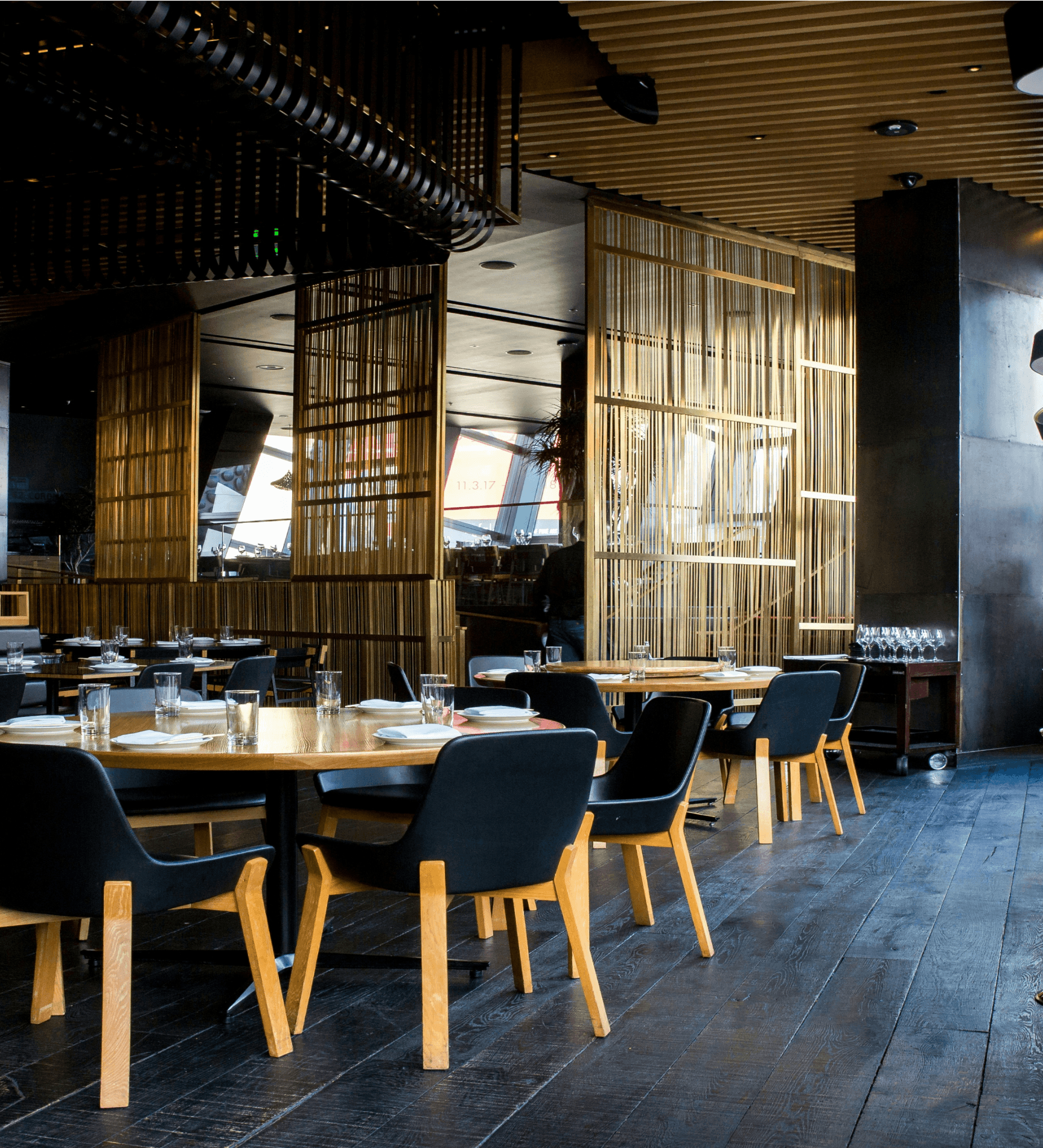 Modern restaurant interior with wooden walls, large windows, and minimalist tables and chairs arranged for dining.