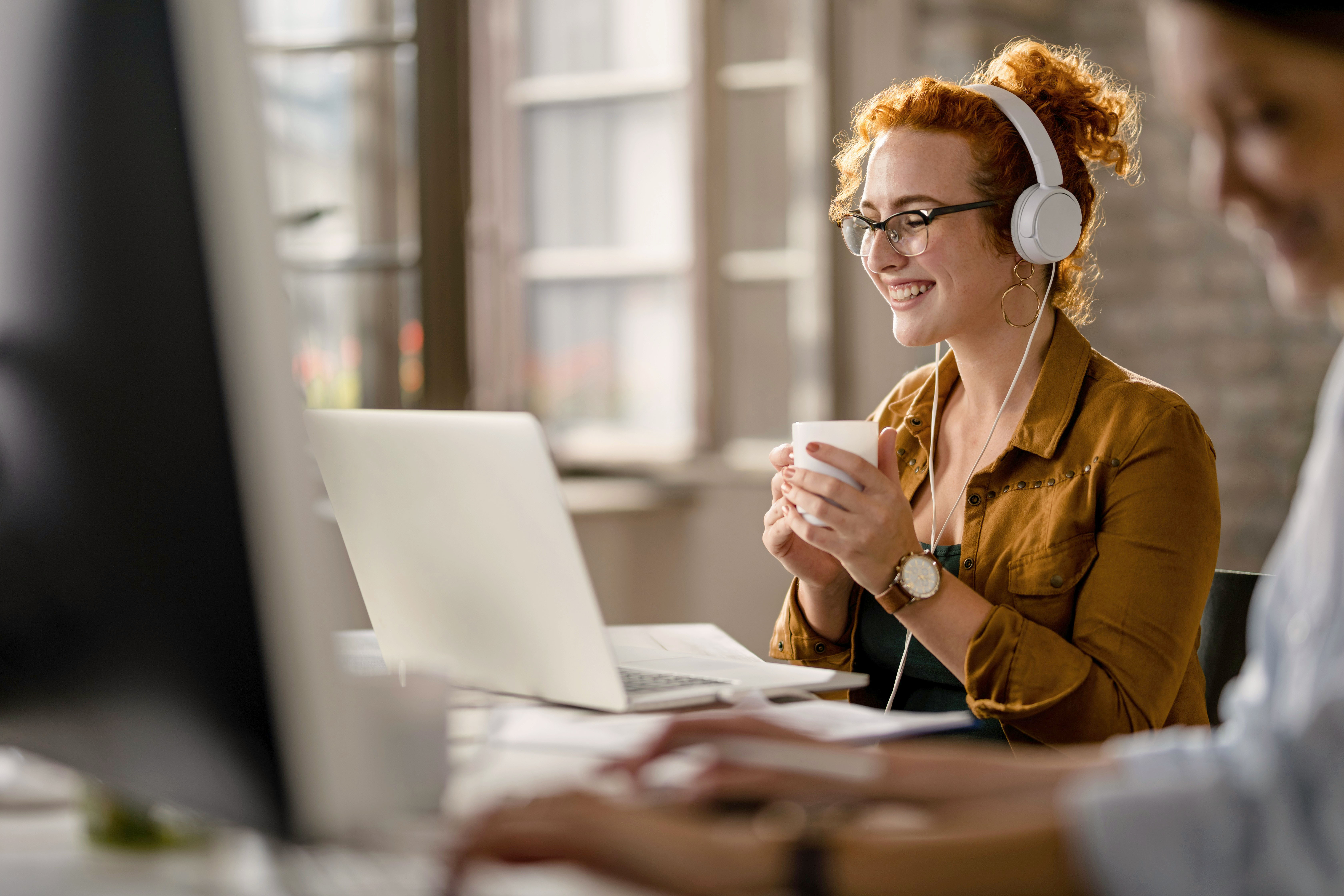 A person wearing headphones works on a laptop, focused, in a well-lit office space. Another person is nearby.