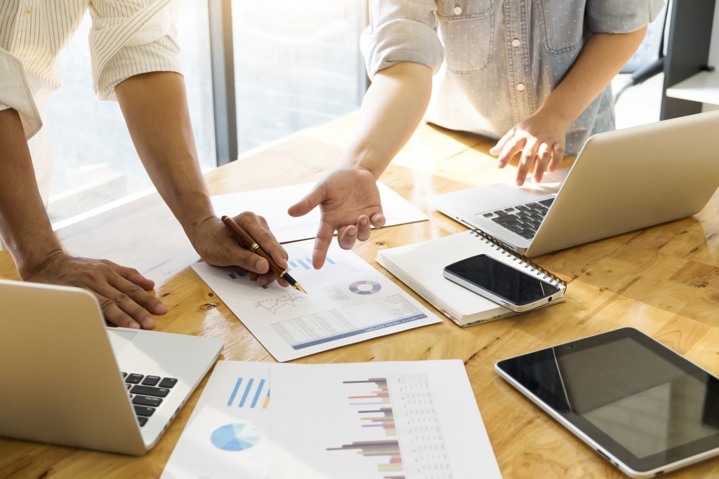 Hands review printed charts on a table with laptops, a phone, and a tablet nearby.