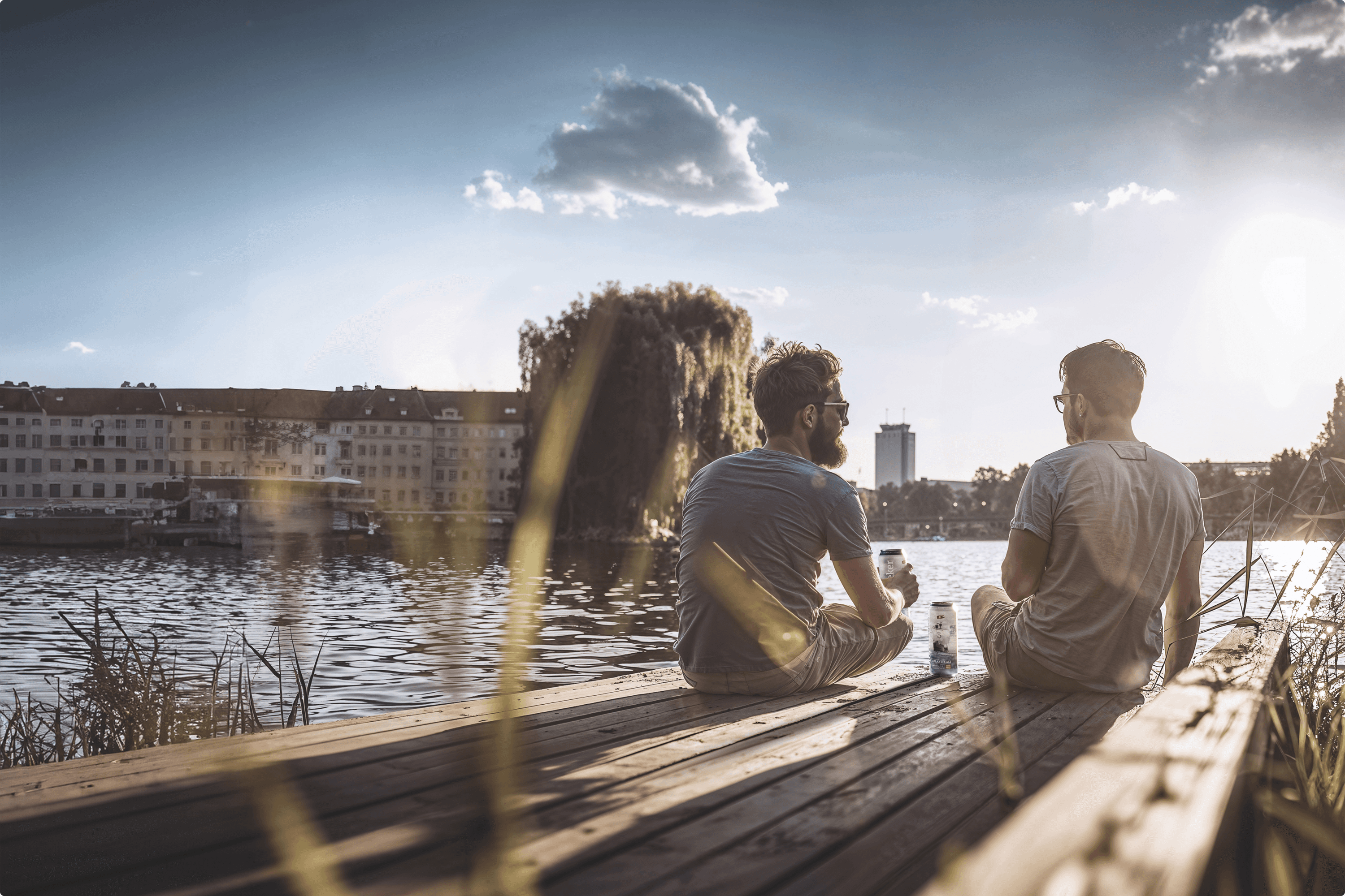Two men sit on a dock with drinks, looking at a lake.