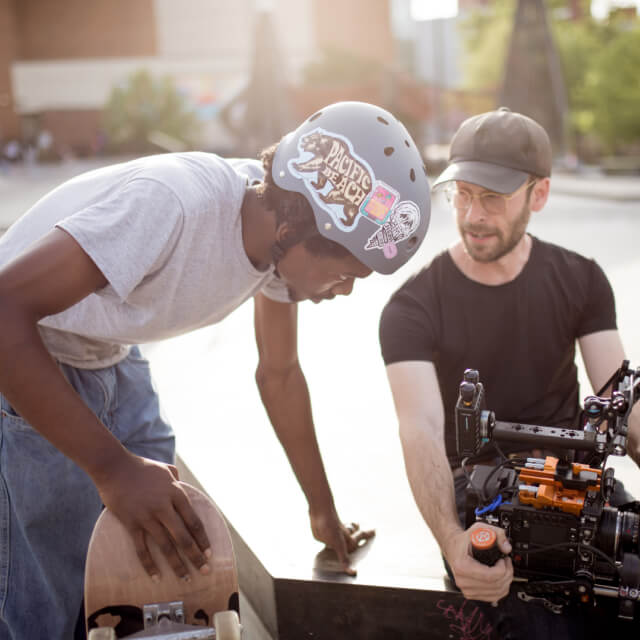 A skateboarder wearing a helmet listens to a man operating a professional film camera outdoors in daylight.