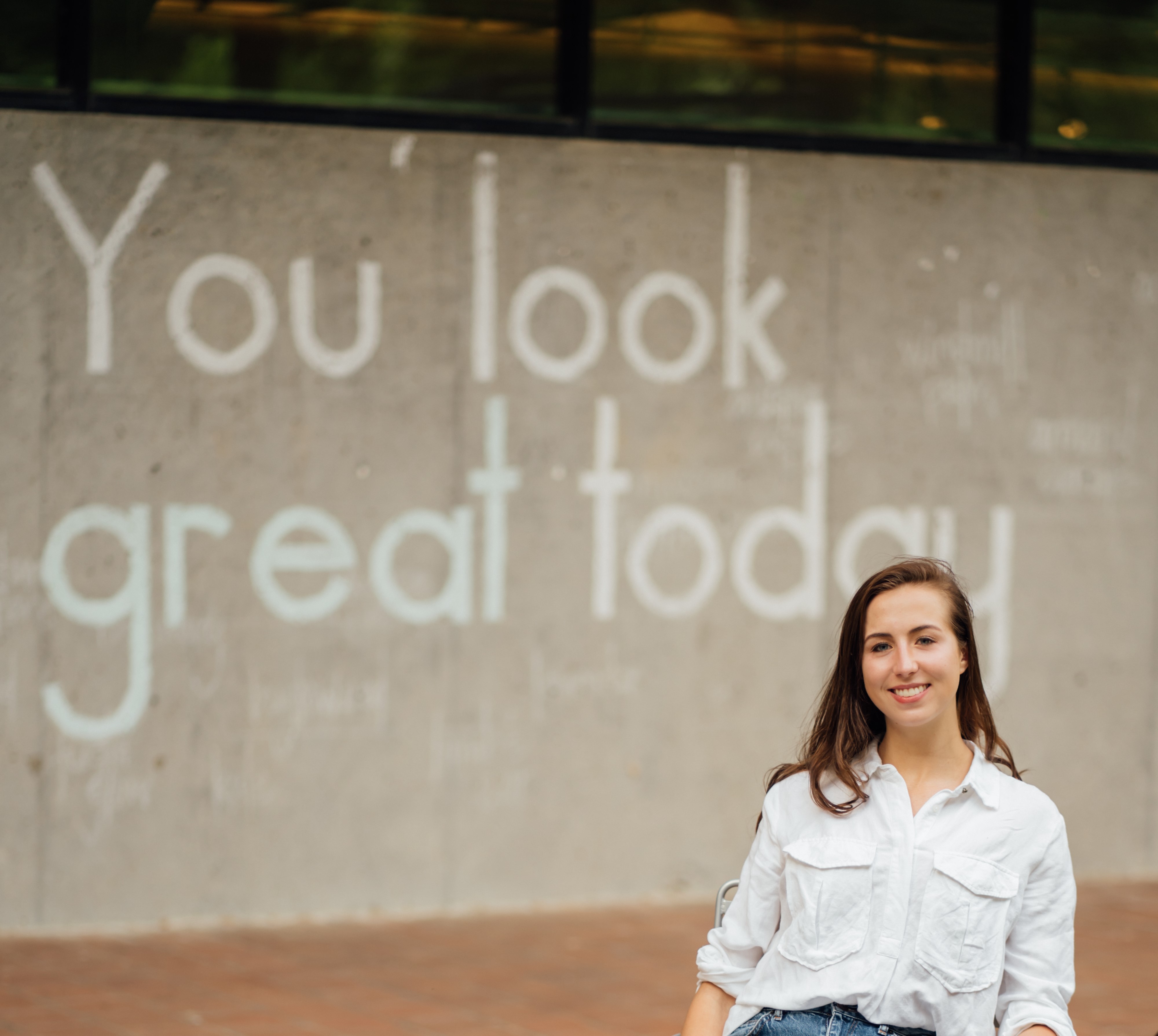 Allie Paschal with a white shirt on in the foreground with a sign that says 'You look great today' in the background