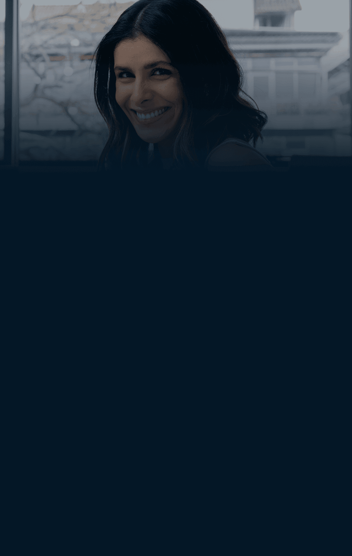 Uma mulher de blusa amarela olha para o celular, sentada perto de uma janela em uma sala suavemente iluminada.