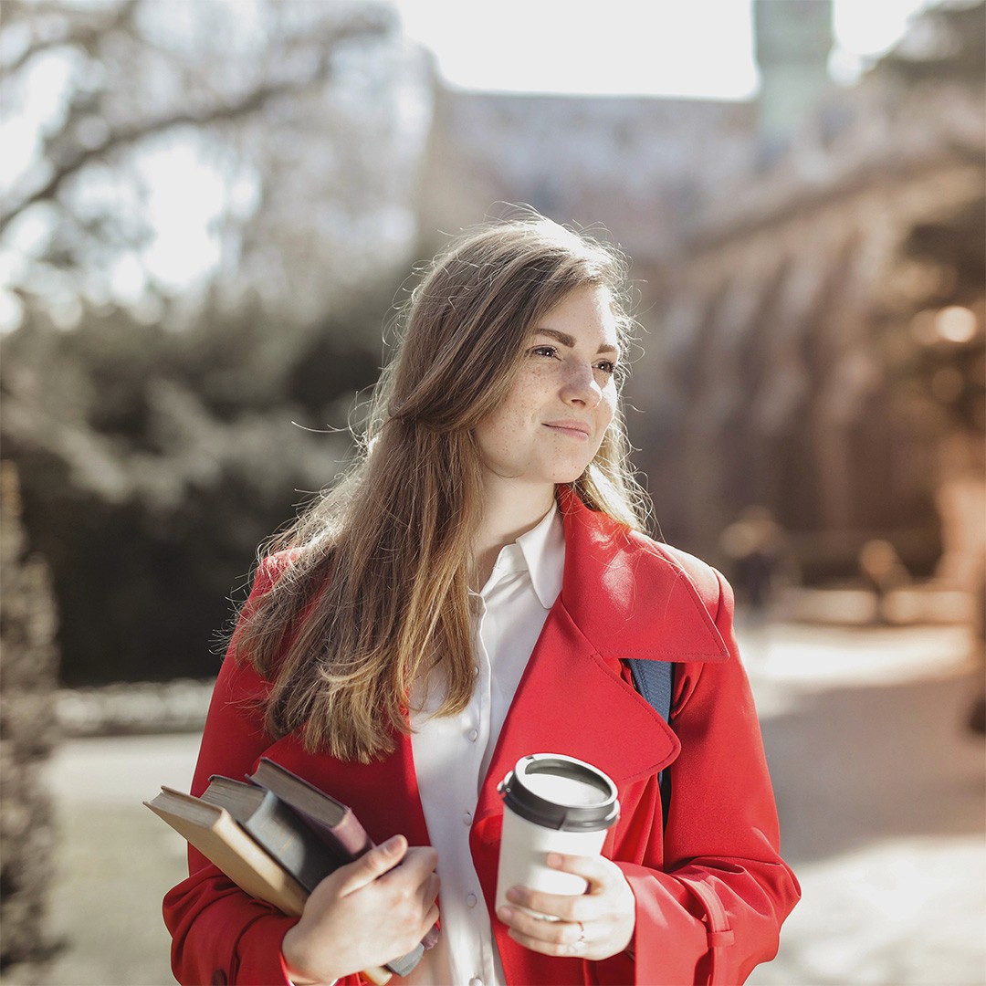 A young girl studient with dark blonde hair wearing a red coat, carries coffee and books across campus, the sun is shining on her face and she is smiling
