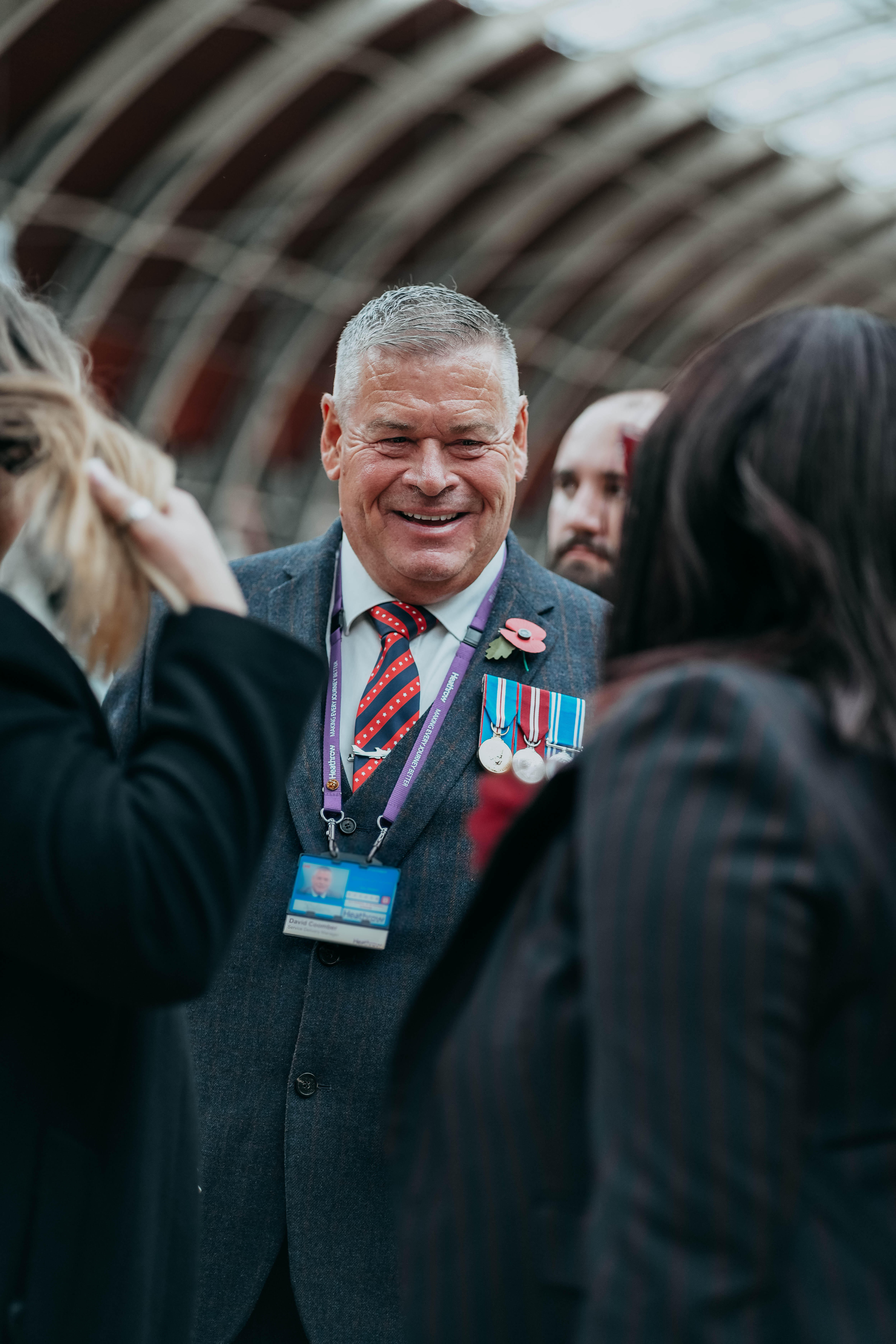 A close-up of a smiling veteran wearing military medals on his suit jacket, engaged in a lively conversation with others inside a train station.