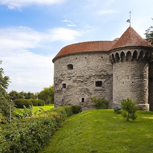 Kővár kerek tornyokkal és vörös cseréptetőkkel, zöld fű és bokrok ölelésében, részben felhős kék ég alatt.