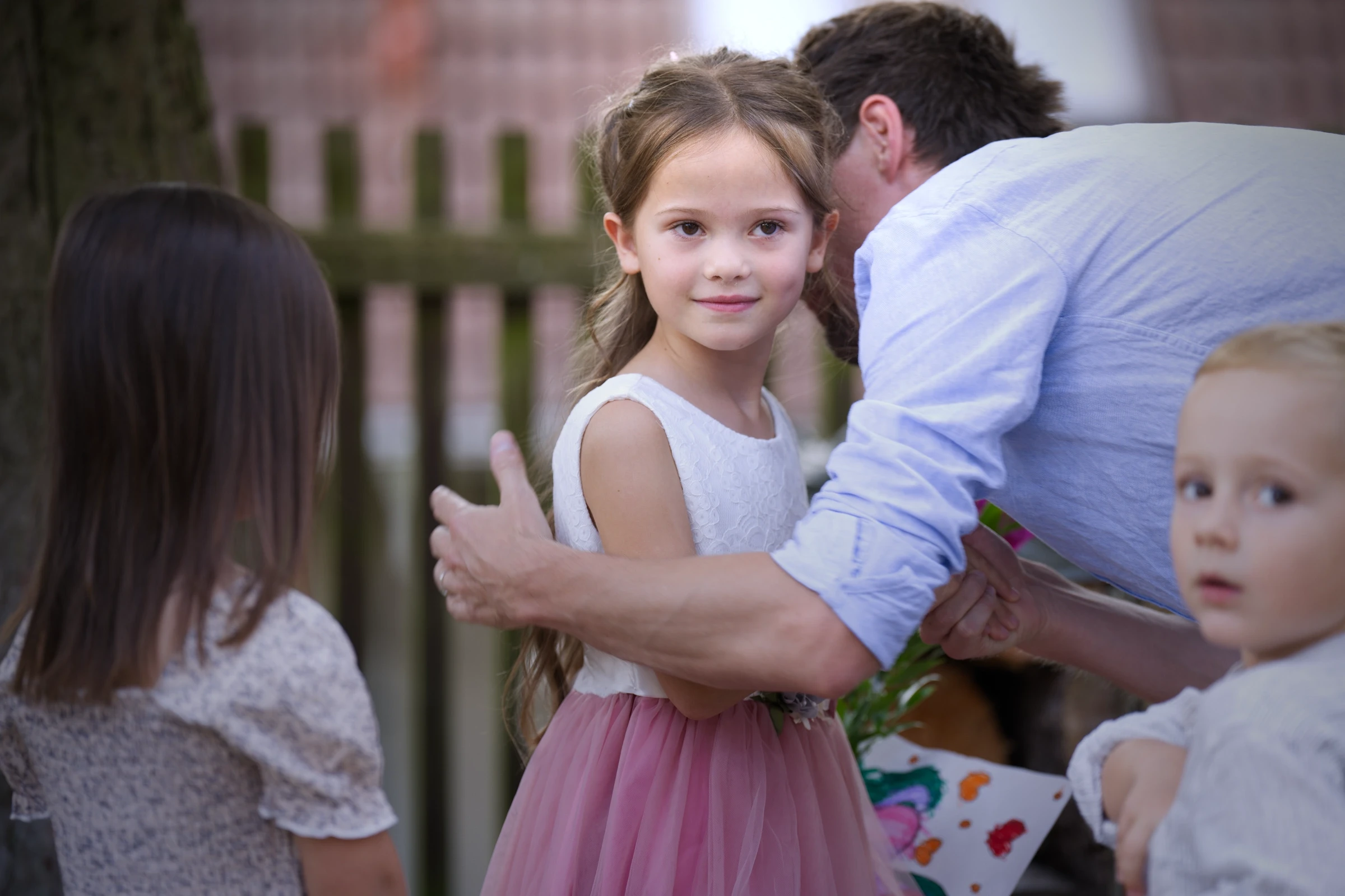 Emmy wird von einem Gast mit Umarmung begrüßt, während sie zur Seite in die Kamera blickt. Um sie herum stehen Geschwister und weitere Kinder, die mit ihr den besonderen Tag der Einschulung gemeinsam erleben.