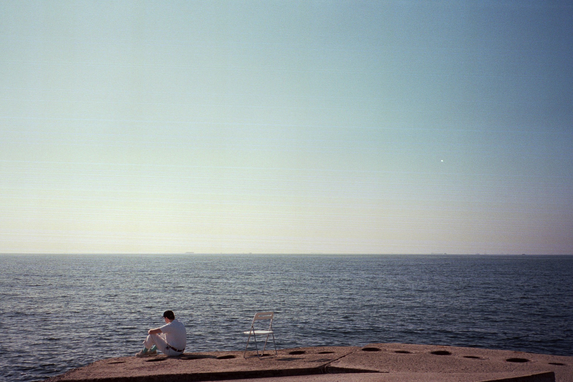A solitary person sits on a concrete pier beside an empty chair, gazing at the vast expanse of the calm sea under a clear blue sky.