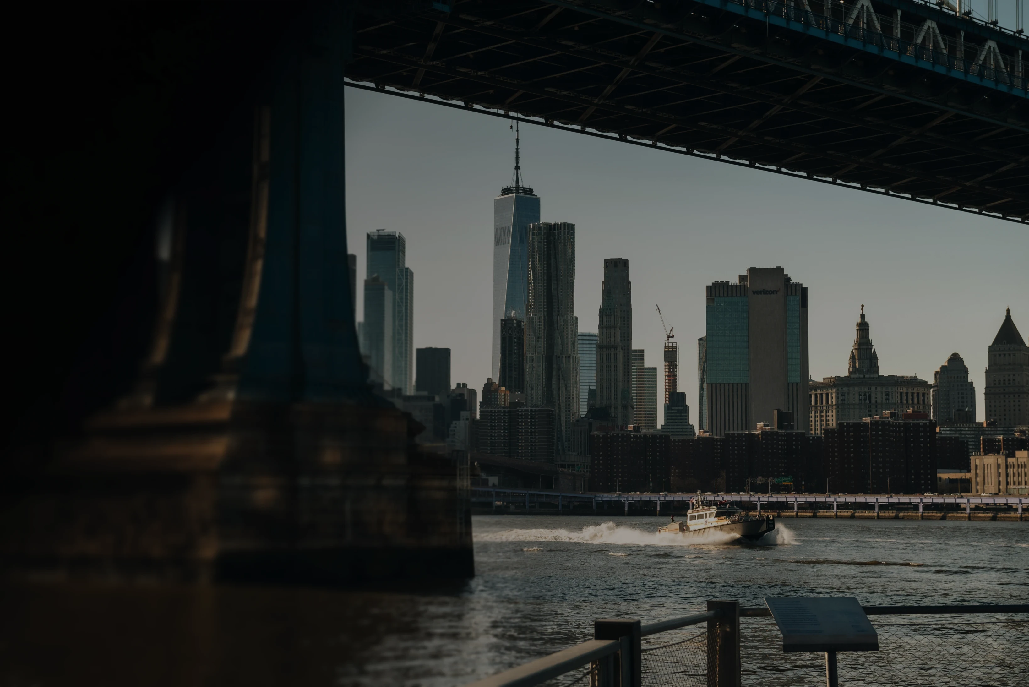 View of the Lower Manhattan skyline and One World Trade Center from beneath the Brooklyn Bridge, with a boat crossing the East River.