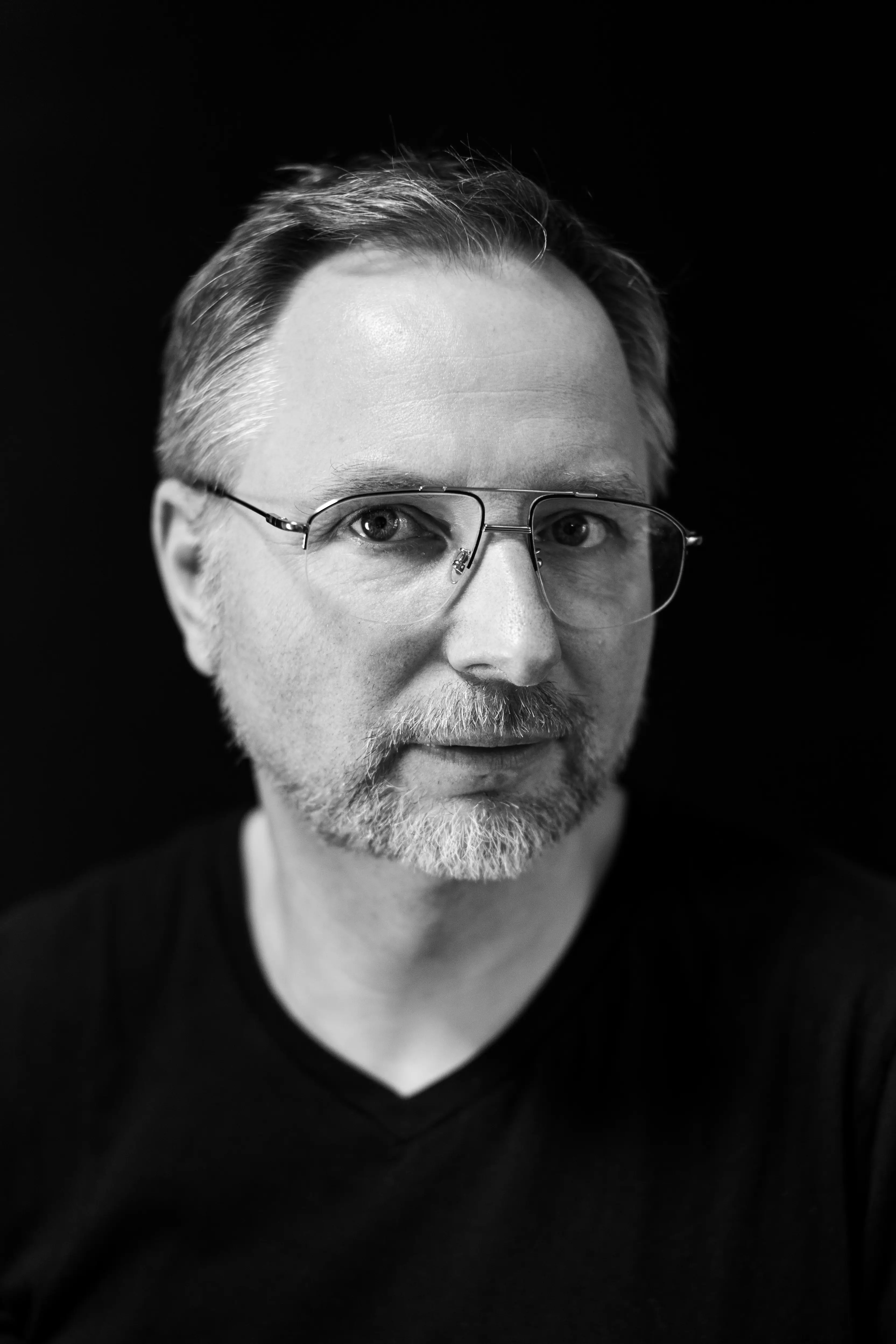 Mężczyzna w okularach z siwą brodą i krótkimi włosami, ubrany w czarną koszulę, na tle czarnym. Patrzy w kamerę z neutralnym wyrazem twarzy.
