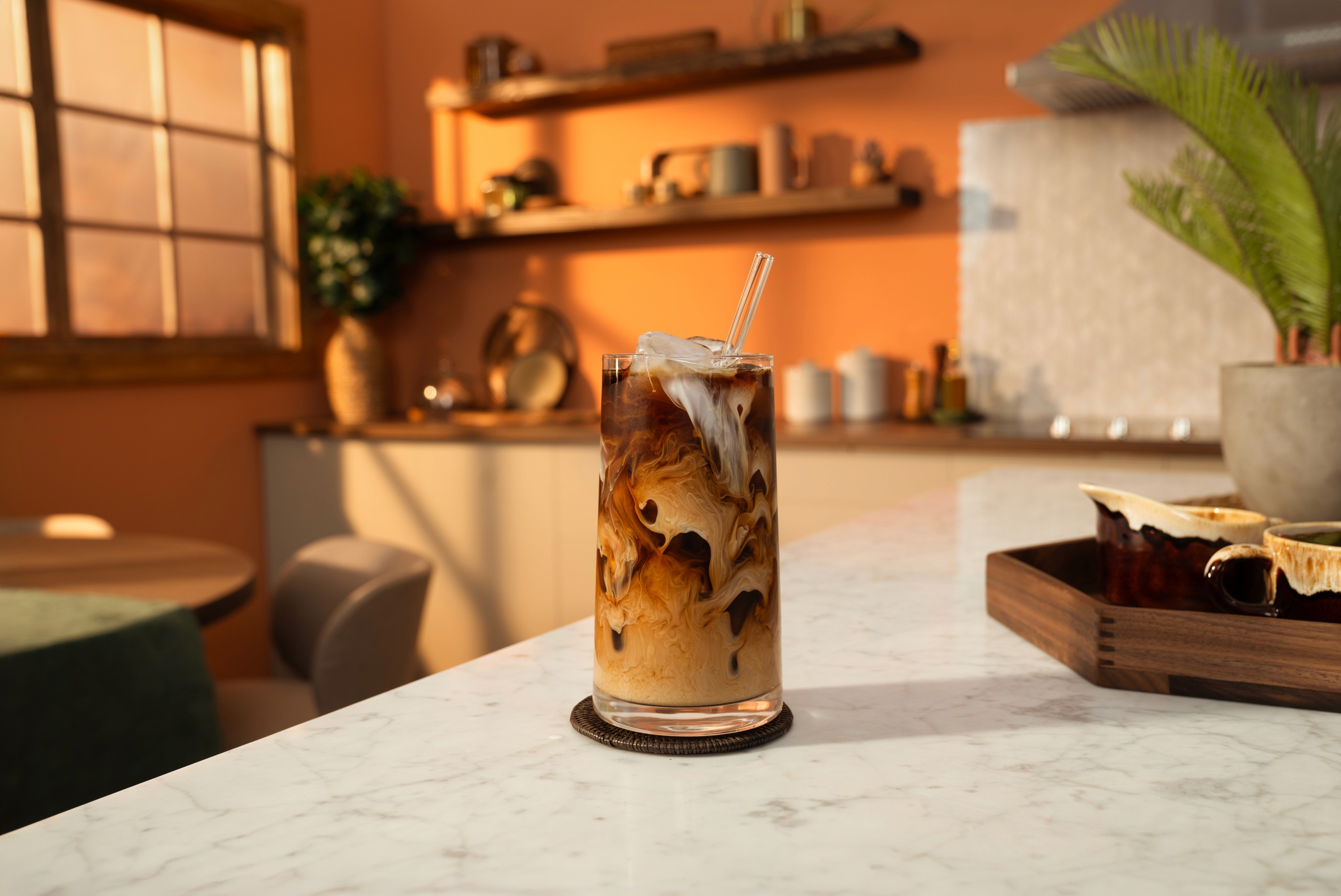 Iced coffee styled in a clear glass with a clear glass straw with cream cascading through the ice and coffee sitting on a home kitchen counter.