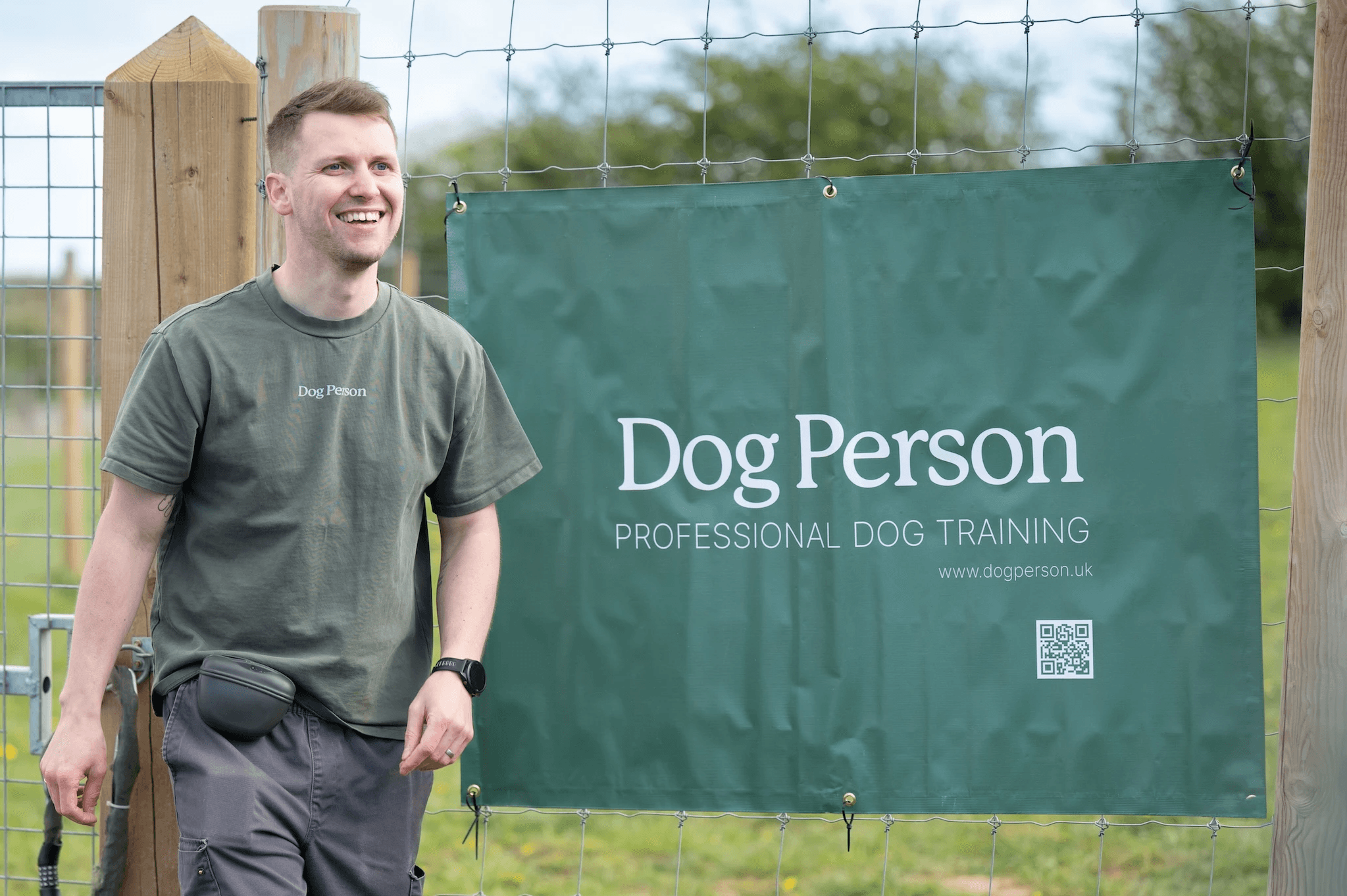 Scott (Dog Person) wearing a Dog Person branded t-shirt, smiling next to a Dog Person branded advertising board
