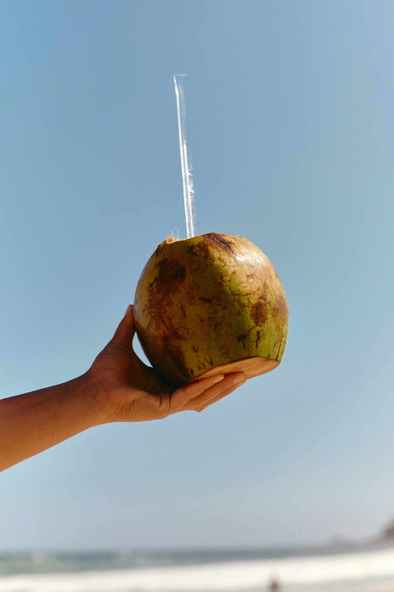 Une main tient une noix de coco verte fraîche avec une paille, sur fond de ciel bleu et de plage floue.