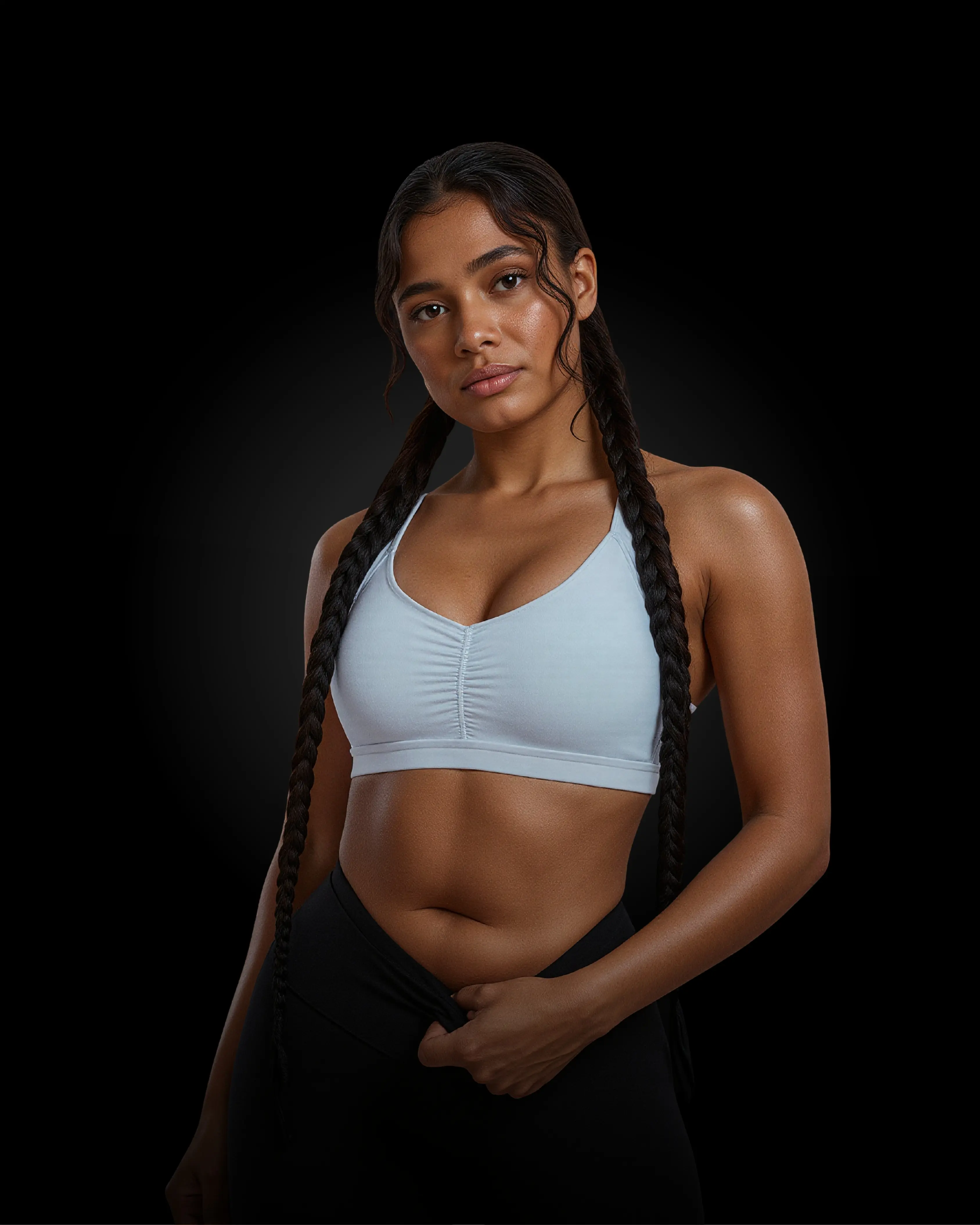 A fit woman with long braids wearing a white sports bra and black leggings. She looks directly at the camera with a confident expression.