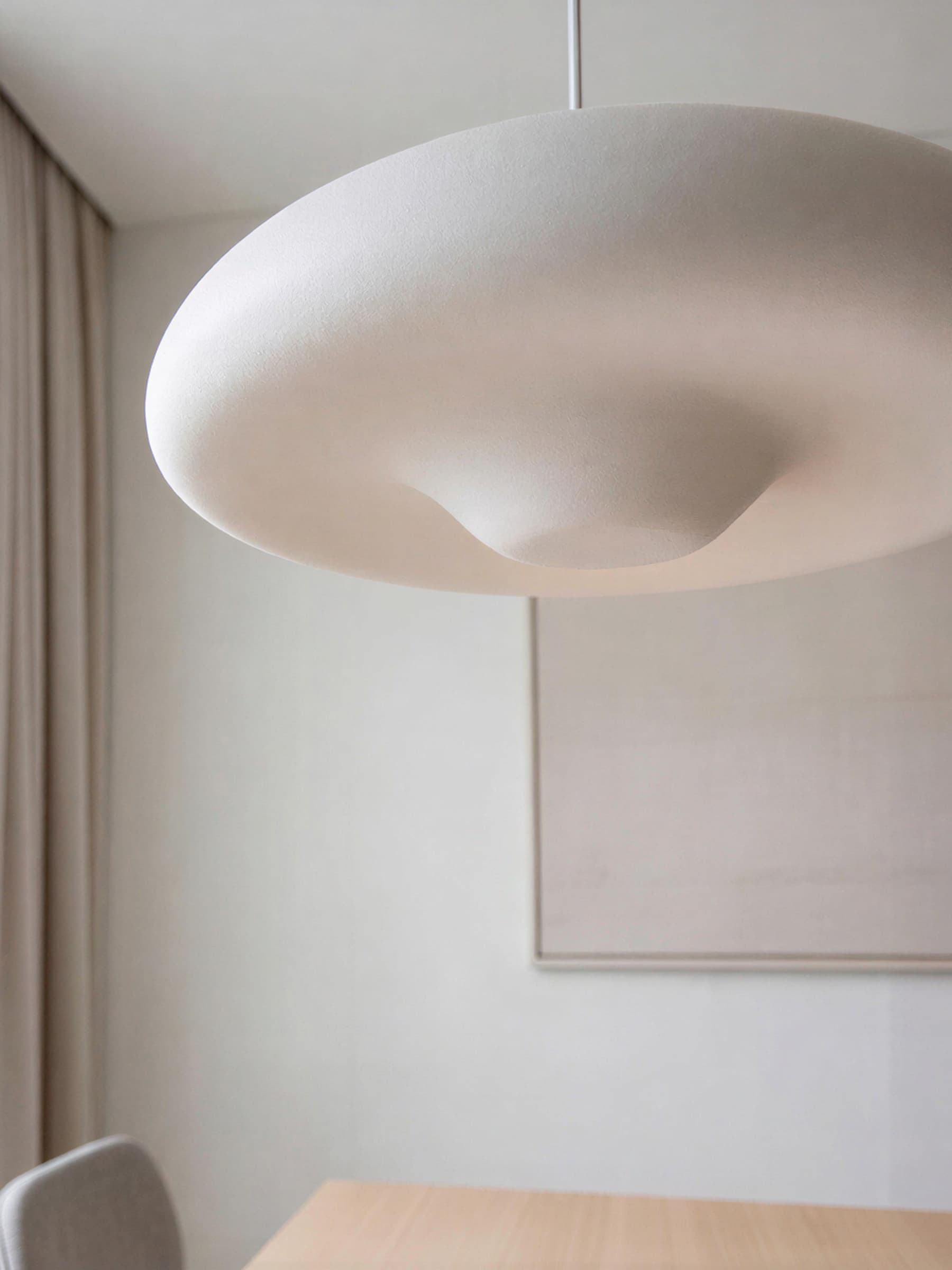 Modern, minimalist interior with a large, dome-shaped white pendant light fixture hanging above a light wooden table, set against neutral-colored walls and soft, flowing curtains.