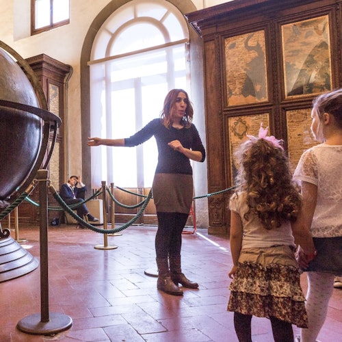 Una donna sta parlando con due bambini in una sala espositiva con un grande globo terrestre e mappe storiche alle pareti.