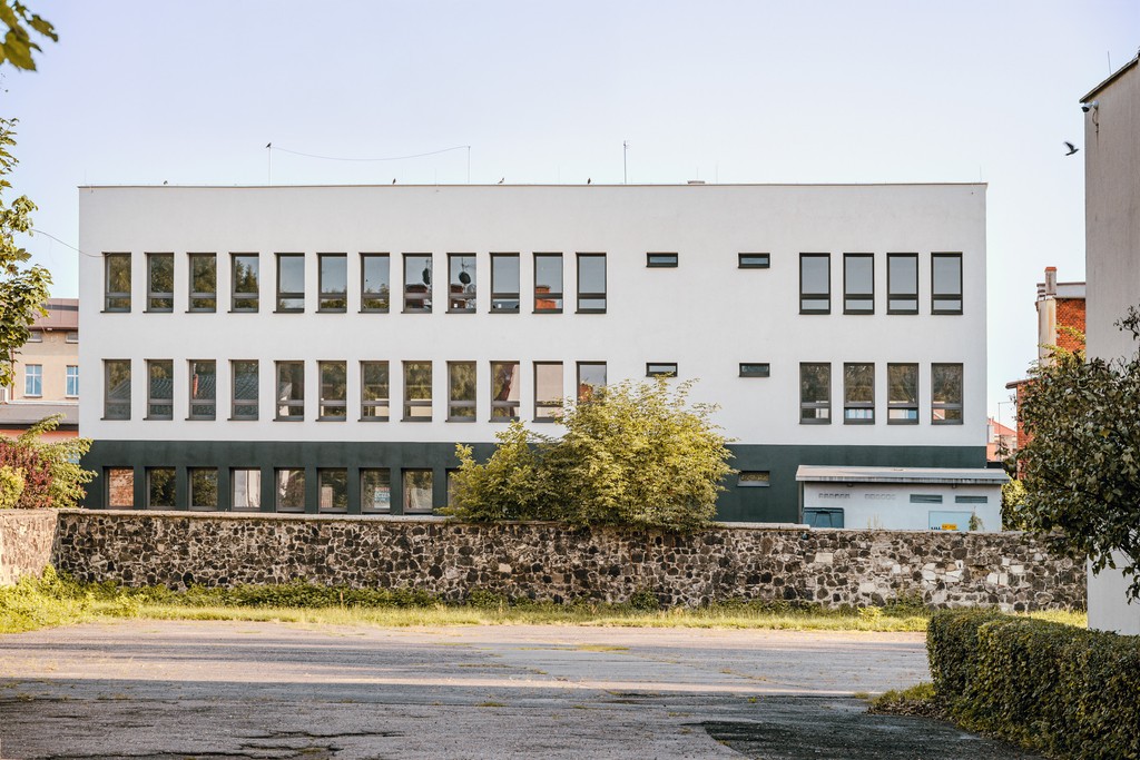 Budynek z wieloma oknami, prawdopodobnie szkoła lub biuro. Zielony pas na dole budynku i kamienny murek.