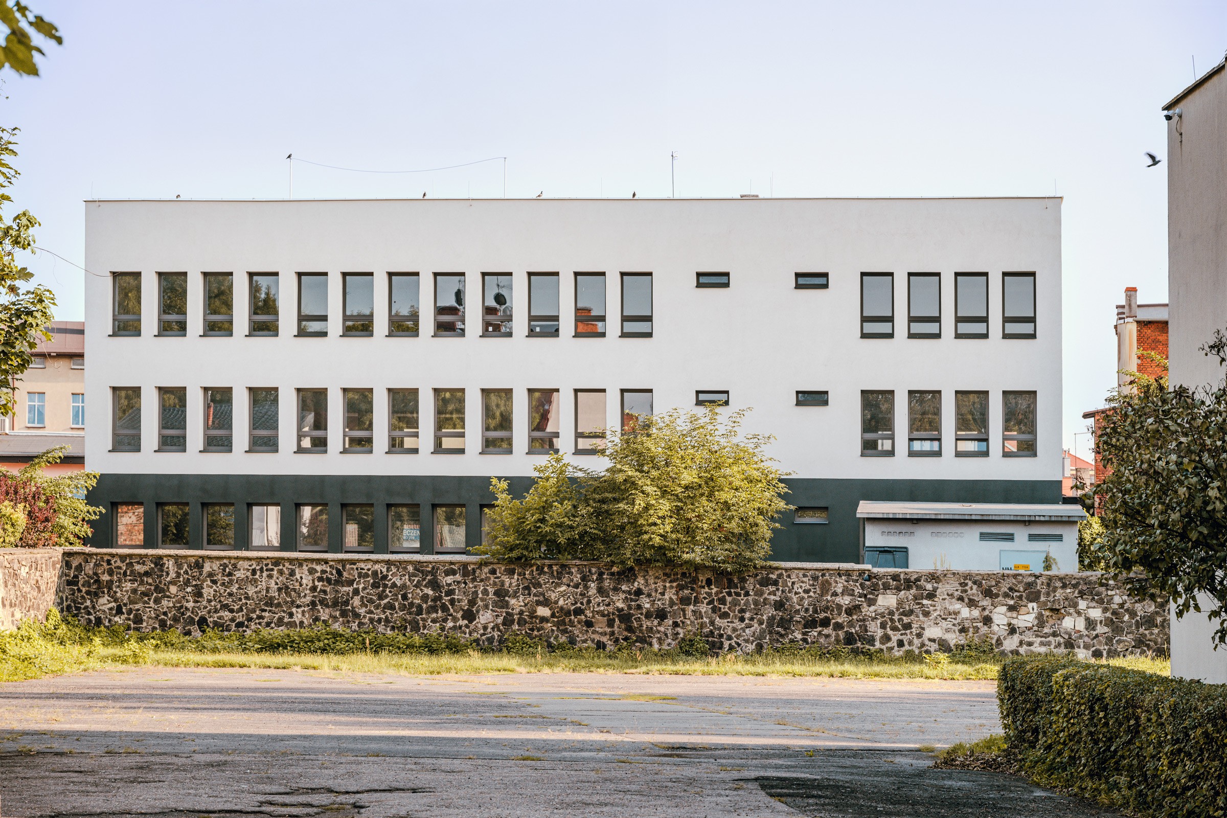 Budynek z wieloma oknami, prawdopodobnie szkoła lub biuro. Zielony pas na dole budynku i kamienny murek.
