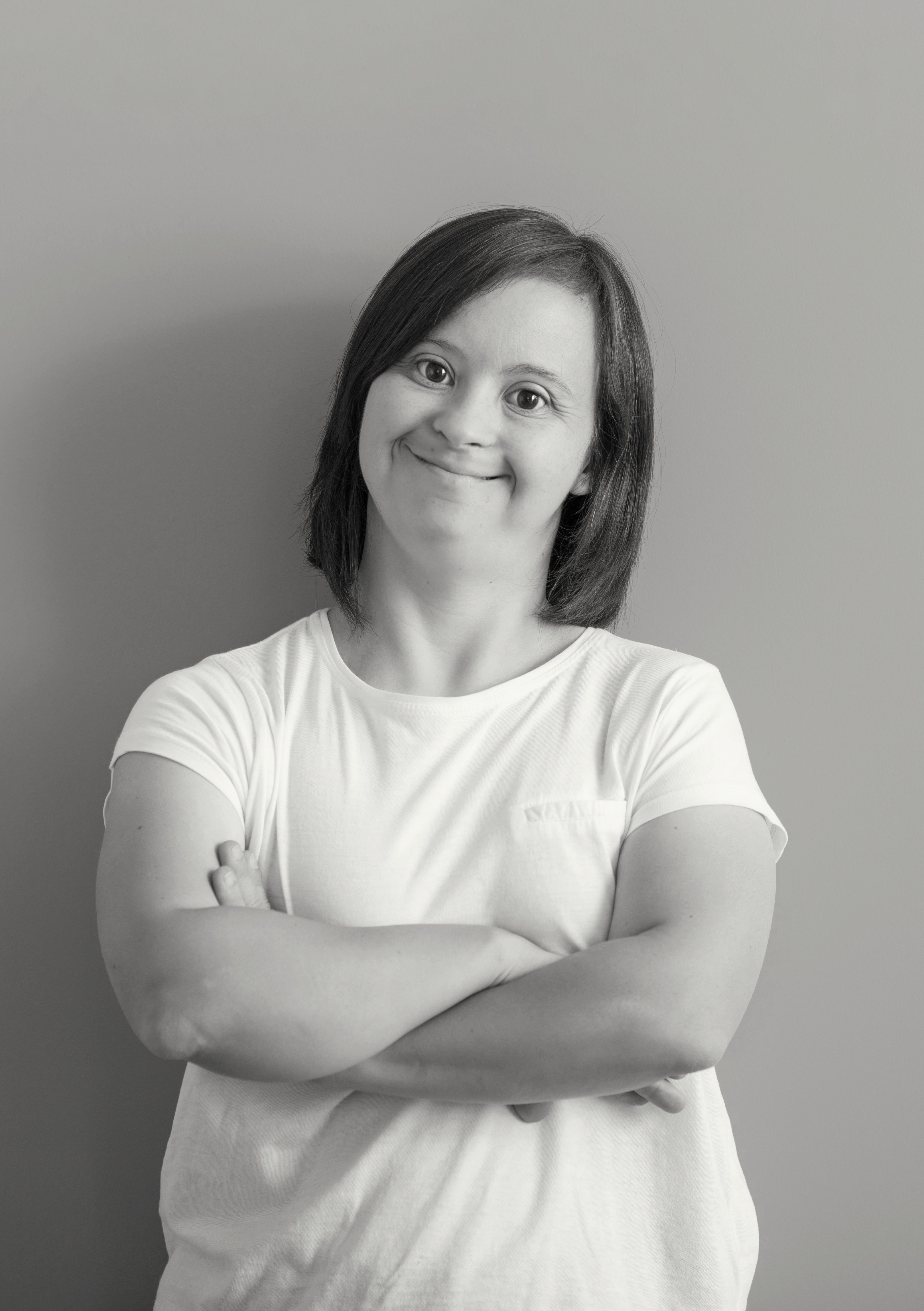 A black-and-white portrait of a smiling young woman with Down syndrome standing with her arms crossed against a plain studio background.