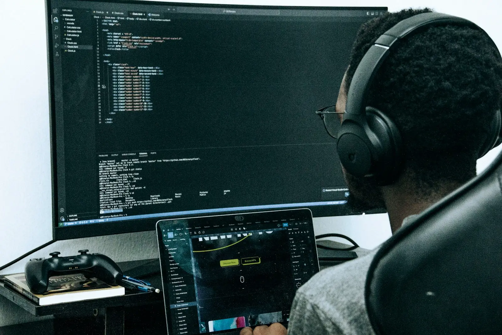 Developer wearing headphones and glasses writing HTML code on a large curved monitor, with a laptop showing a design tool and a game controller on the desk