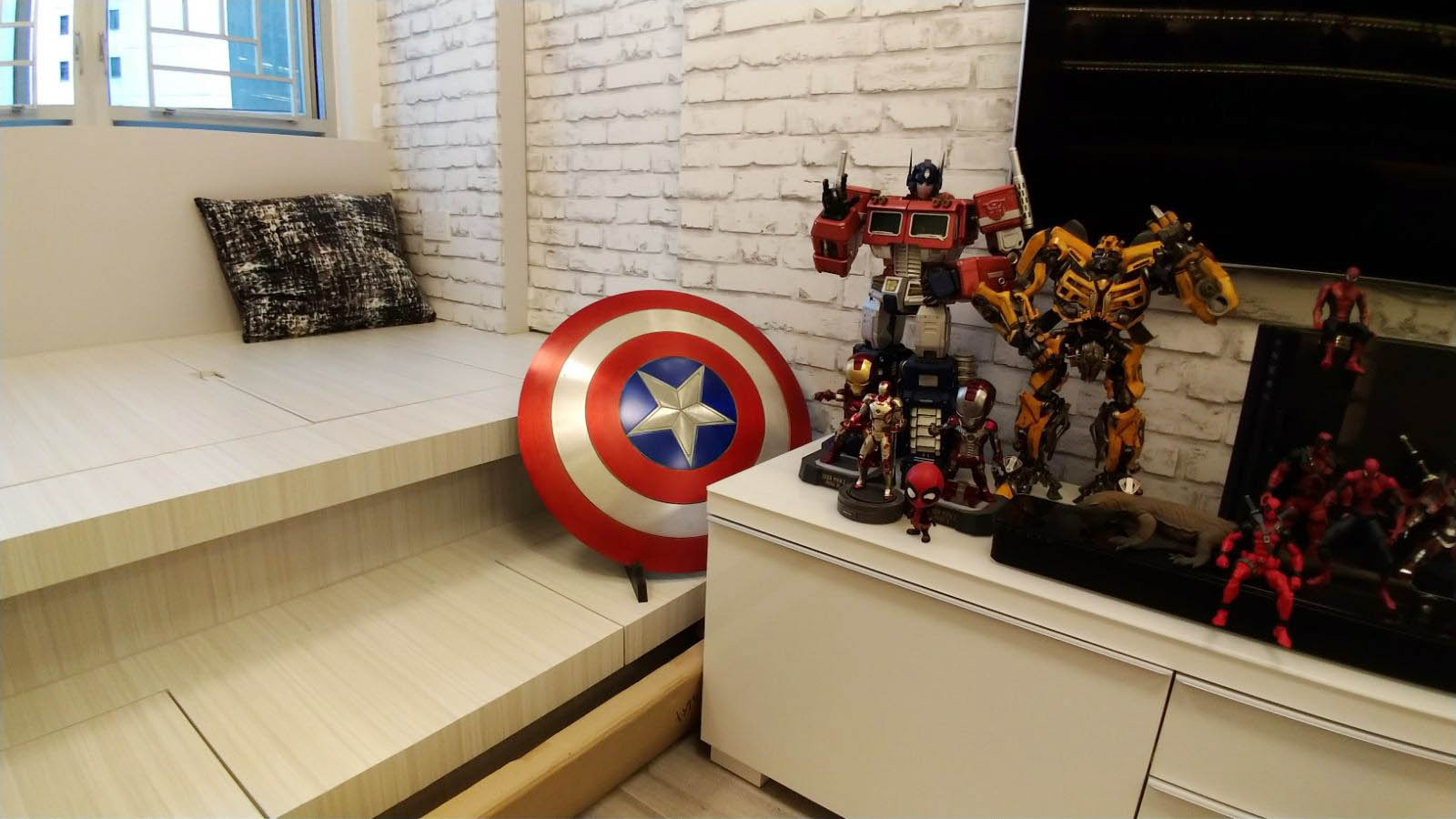 Based on the image provided, here are the details regarding the room decoration:Captain America Shield: A large, iconic red, white, and blue shield is prominently displayed against a white brick wall.Transformers Figures: The room features substantial Transformers figures, including Optimus Prime and Bumblebee, positioned on a white shelf.Marvel Collectibles: The display area includes various Marvel action figures, such as Iron Man, Spider-Man, and Deadpool.Interior Design: The space is designed with a specific theme in mind, featuring elevated wooden flooring (tatami-style) and a brick-patterned accent wall to create a structured hobby room