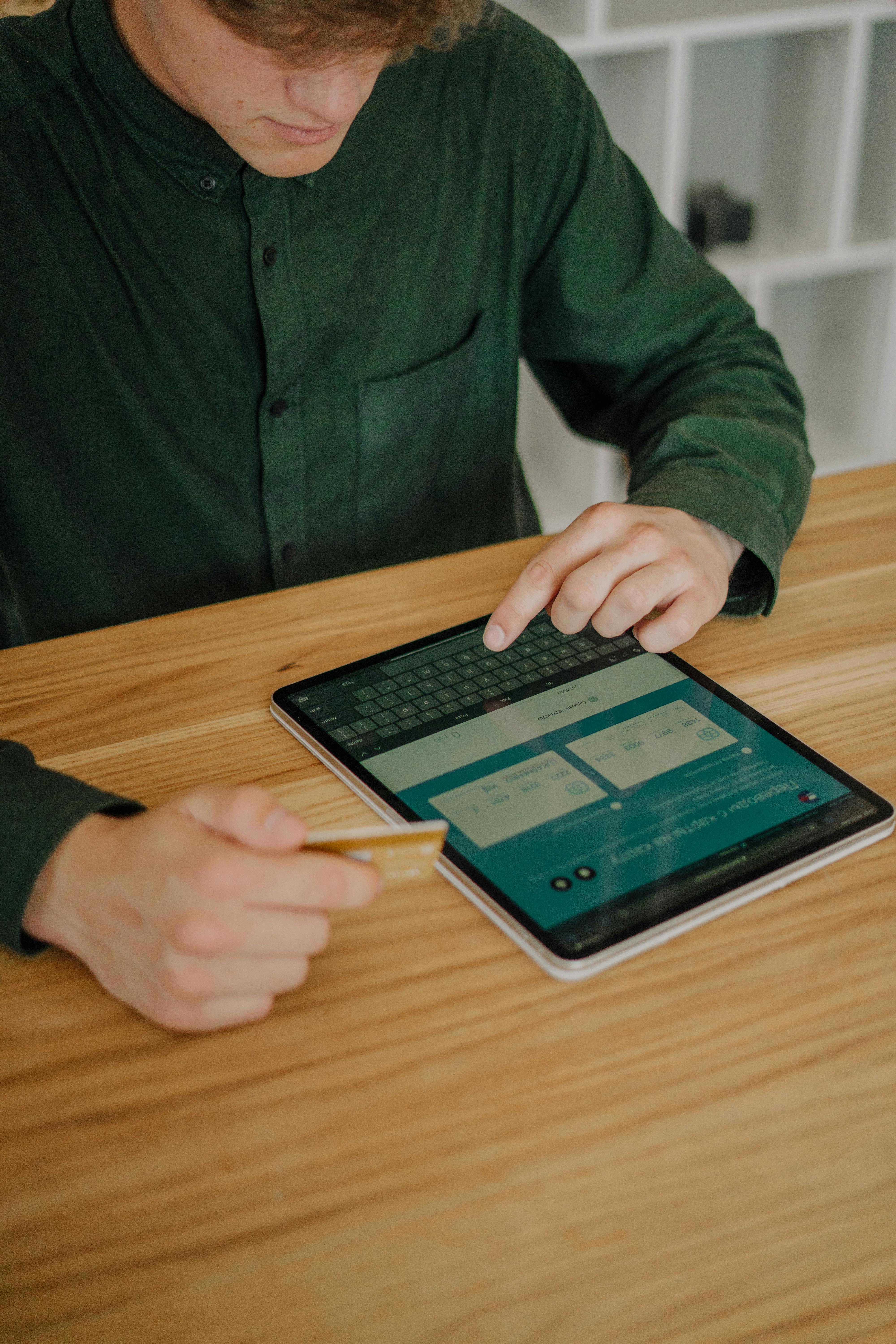 A person in a green shirt holds a credit card while interacting with a tablet displaying online transaction options on a wooden table.