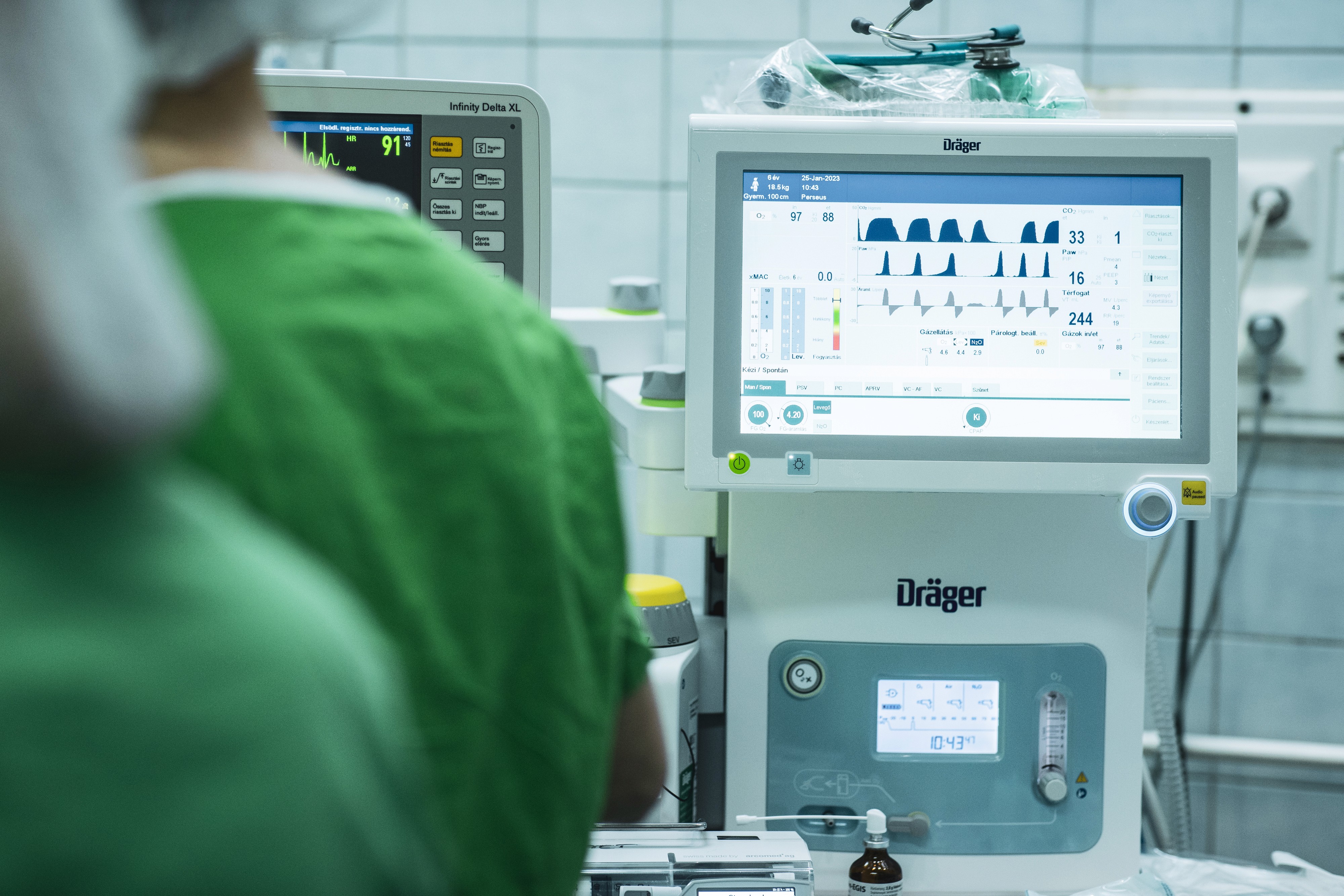 Modern altatógép és monitor orvosi műtőben, háttérben egészségügyi személyzet zöld ruhában.