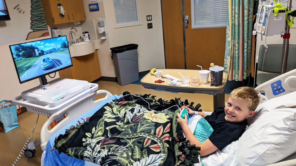 Patient holding an Xbox controller while smiling and looking at the camera. Patient is in a hospital bed with a GO Kart at the end of the bed and Forza playing on the screen.