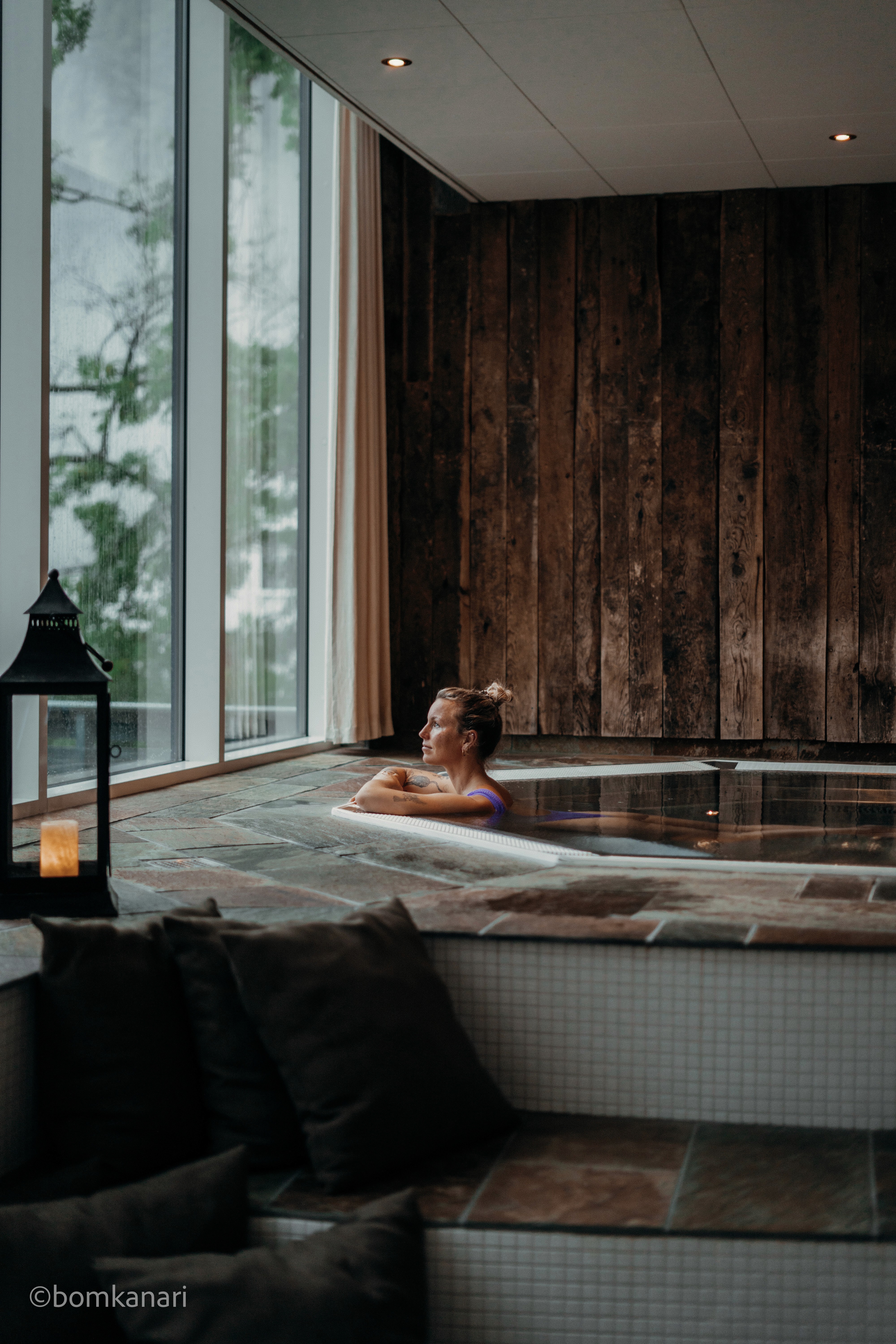 A woman in a white robe sits on a bench, while another person stands in a bright, serene spa setting.