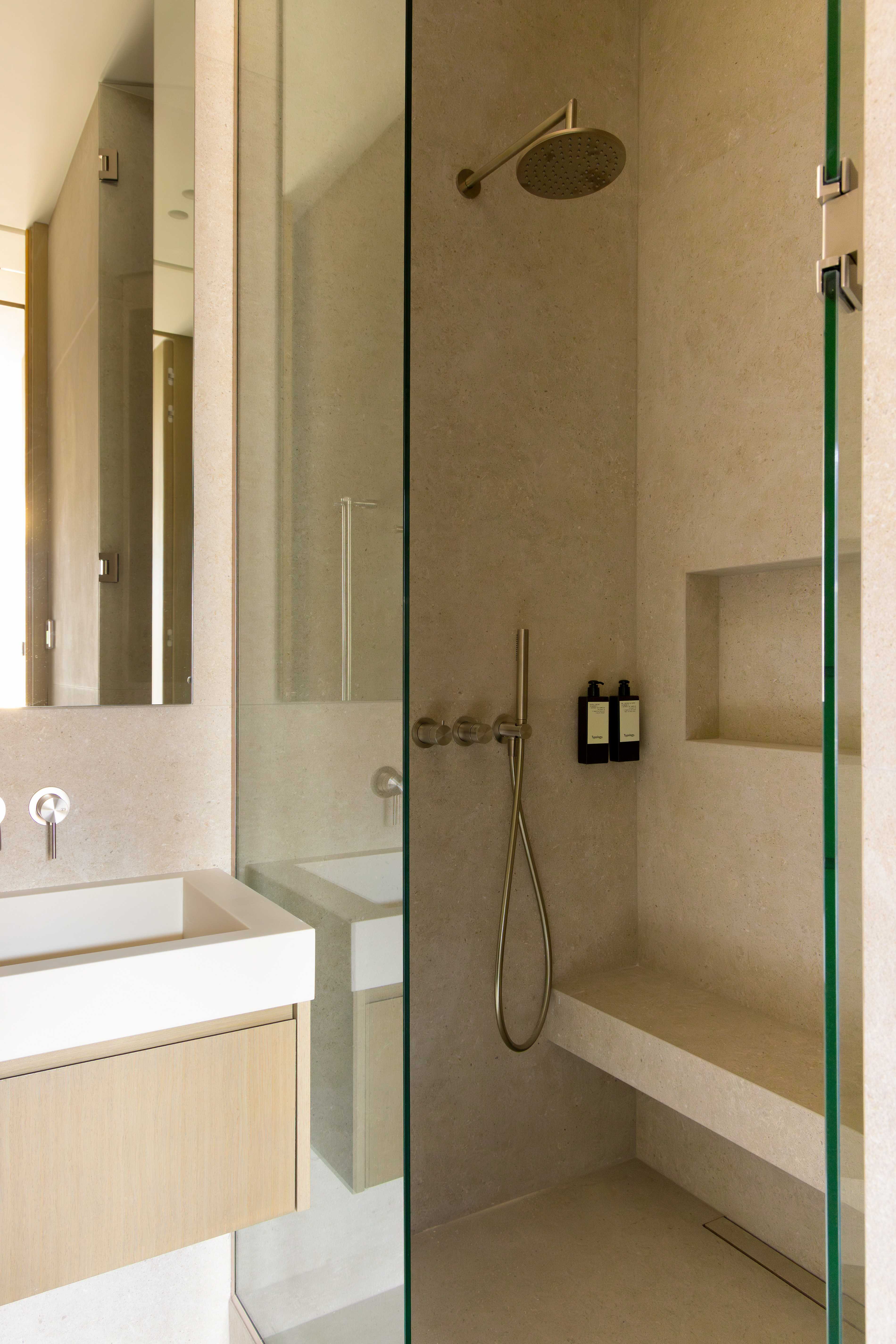 Salle de bains avec douche italienne en pierre beige, paroi vitree, vasque suspendue et robinetterie doree