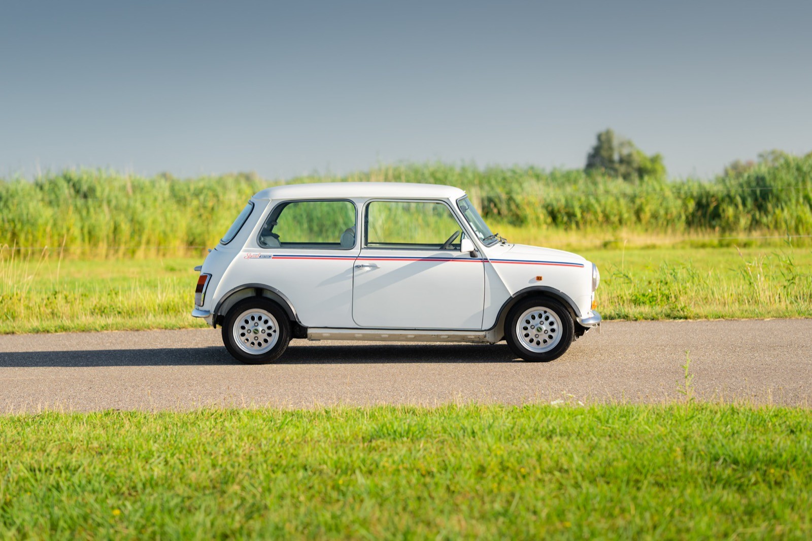 A small white car drives along a road surrounded by lush green fields and a clear blue sky.