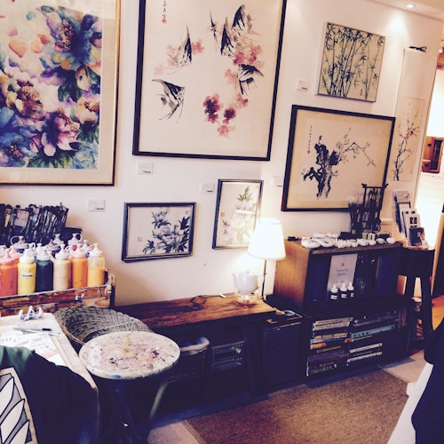 一个舒适的艺术工作室,展示着框装的花卉和鸟类画作,还有一个放置艺术用品的架子和桌子上的小台灯。