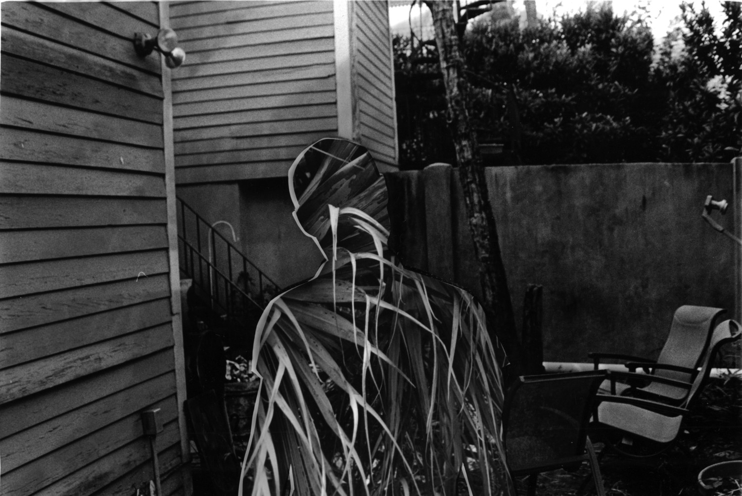 Black-and-white darkroom photograph featuring a silhouetted human figure overlaid with abstract painted textures in an outdoor residential setting, blending photography and mixed media to explore identity, presence, and layered visual narratives.