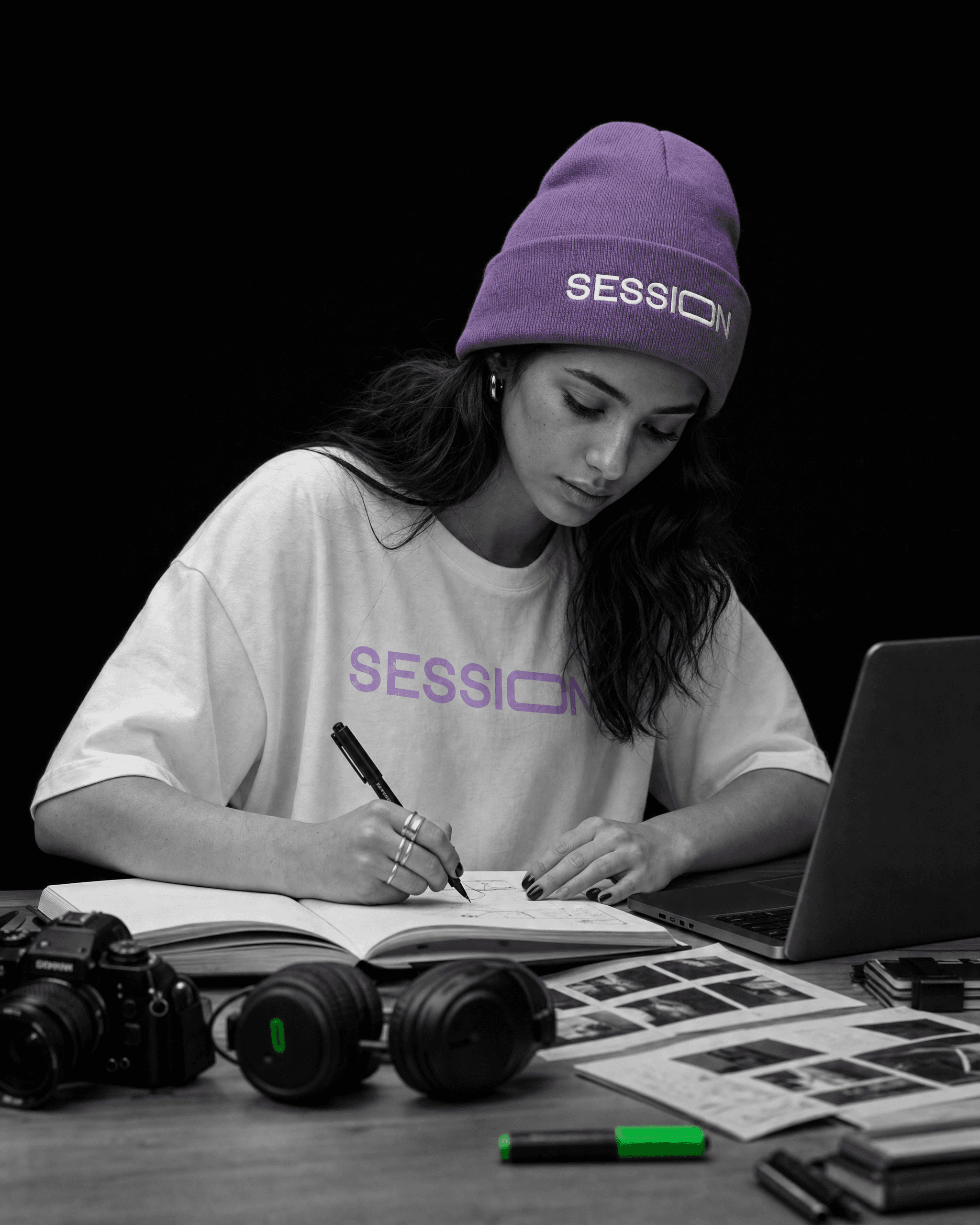 Woman in a light-purple Session Zone beanie and white logo T-shirt writing beside a laptop and camera.