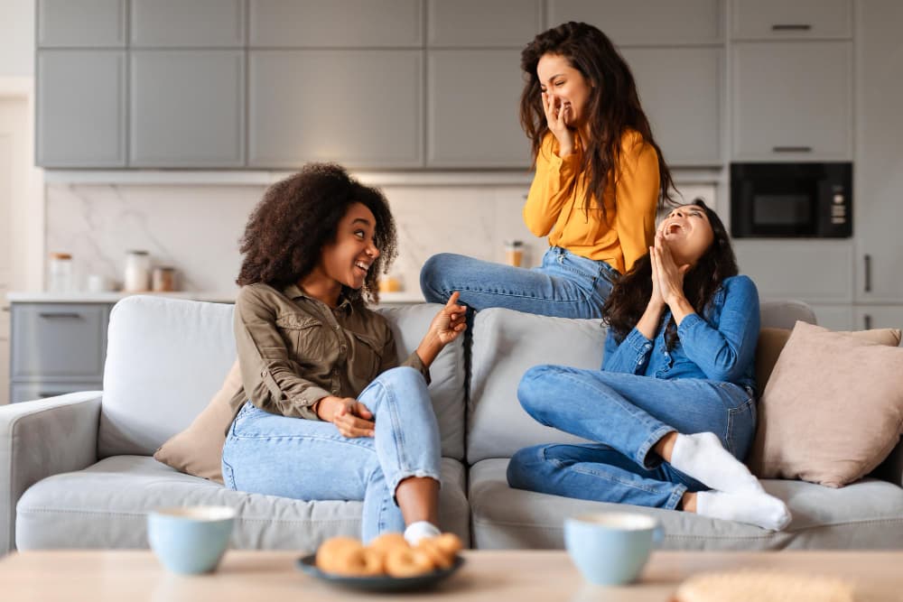 Three women sit and laugh together on a living room couch, enjoying coffee and conversation in a bright, modern home. The image conveys connection, comfort, and the sense of stability that comes with homeownership through Chris Lewis Home Loans.