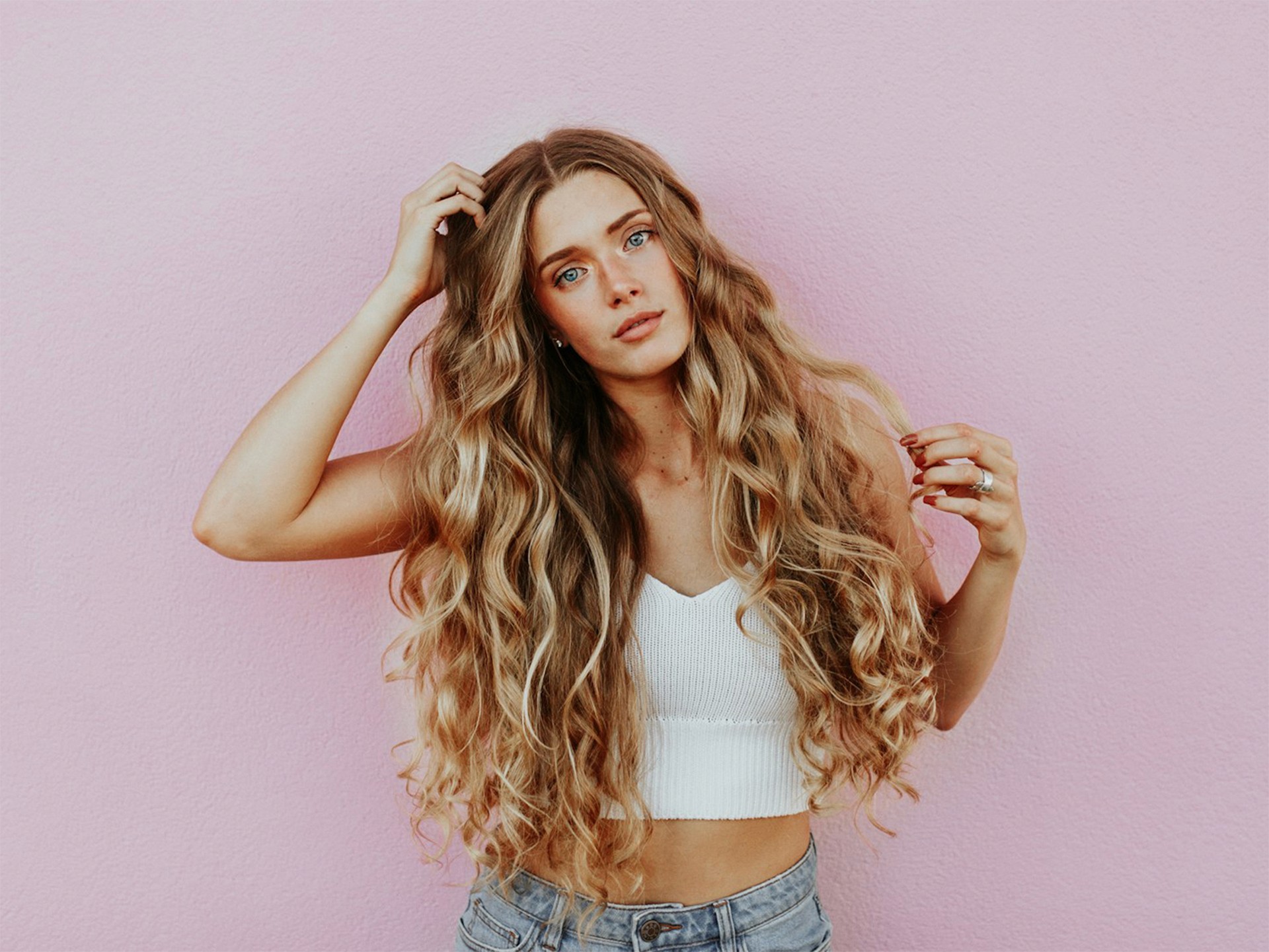 A young woman with long, curly blonde hair poses against a pink wall, holding her hair up with both hands.