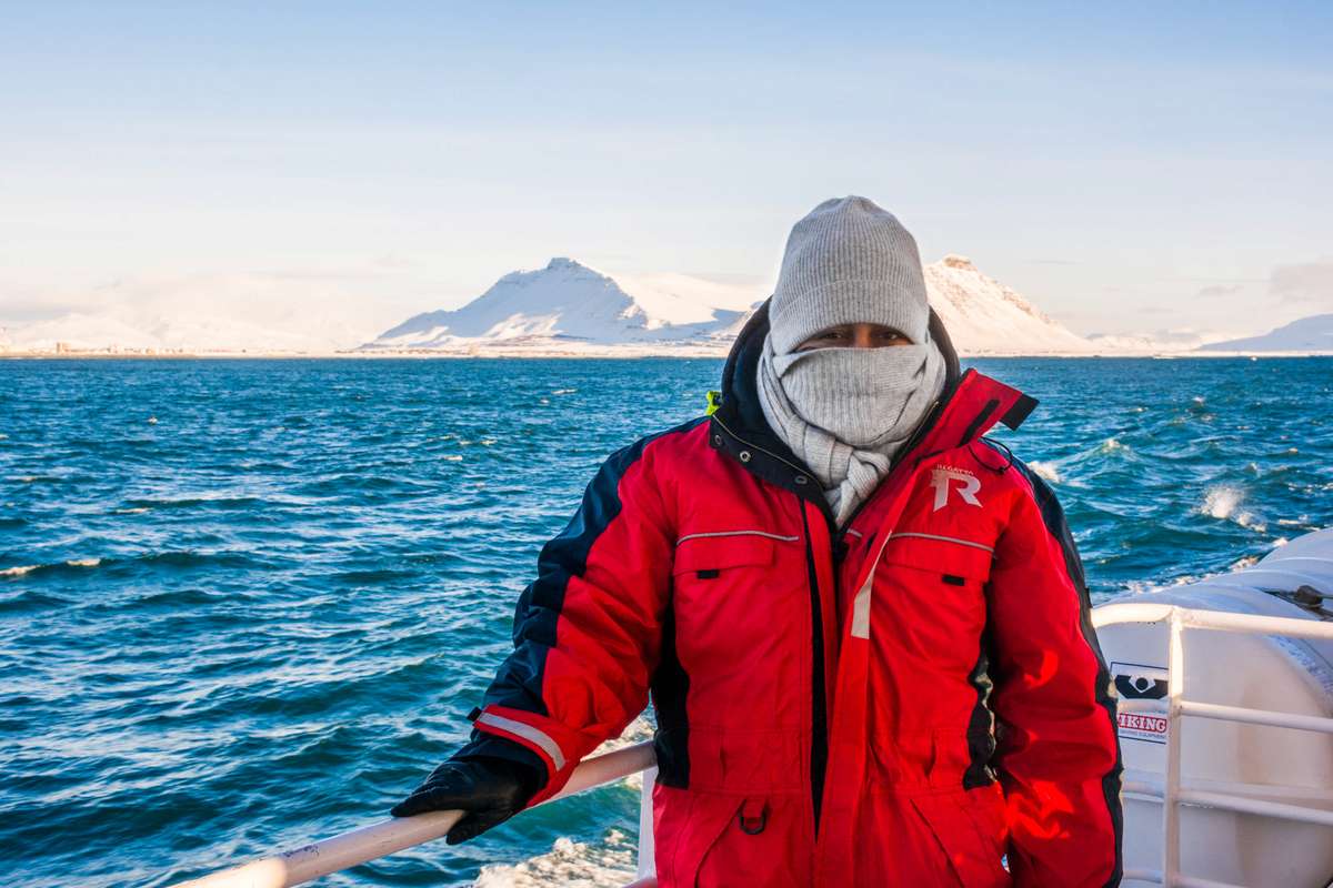 A person in a red and black parka and a white knit balaclava stands on a boat in front of icy mountains and blue water.